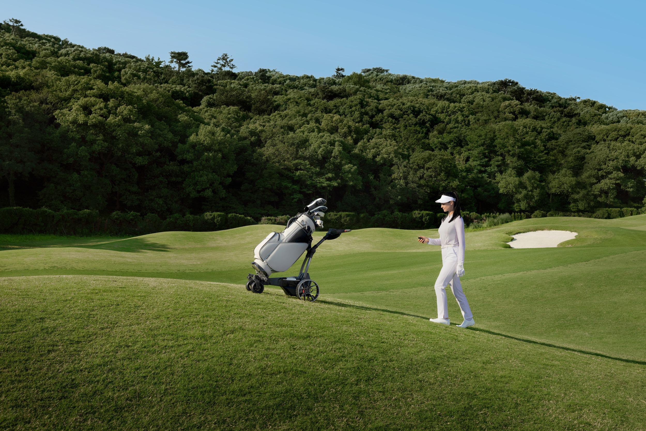 Eine Frau in weißer Kleidung geht neben einem Golfwagen auf einem hügeligen Golfplatz mit Bäumen im Hintergrund.