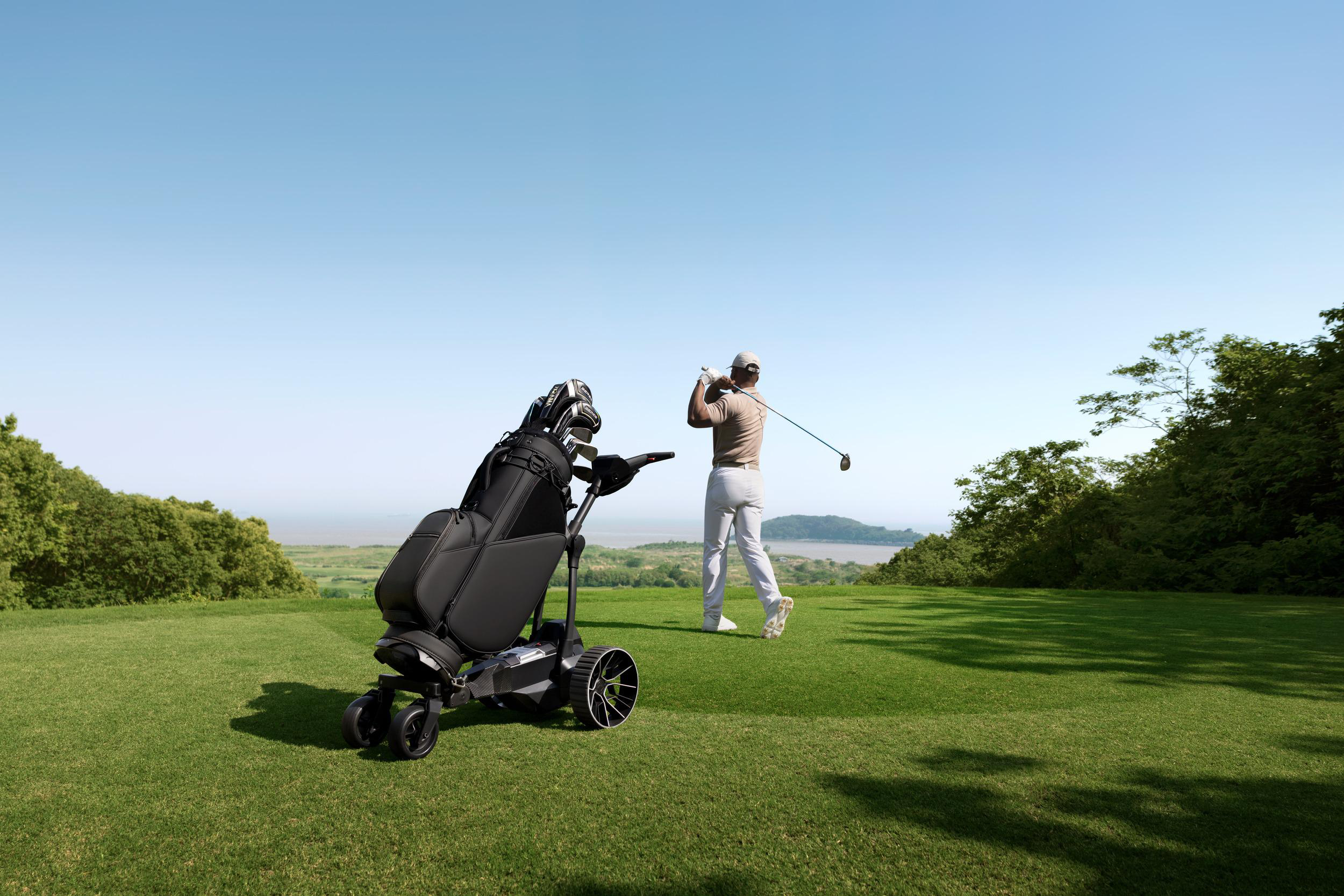 Ein Golfer schlägt auf einem grünen Hügel mit Blick auf das Wasser ab, davor steht ein Golfwagen.