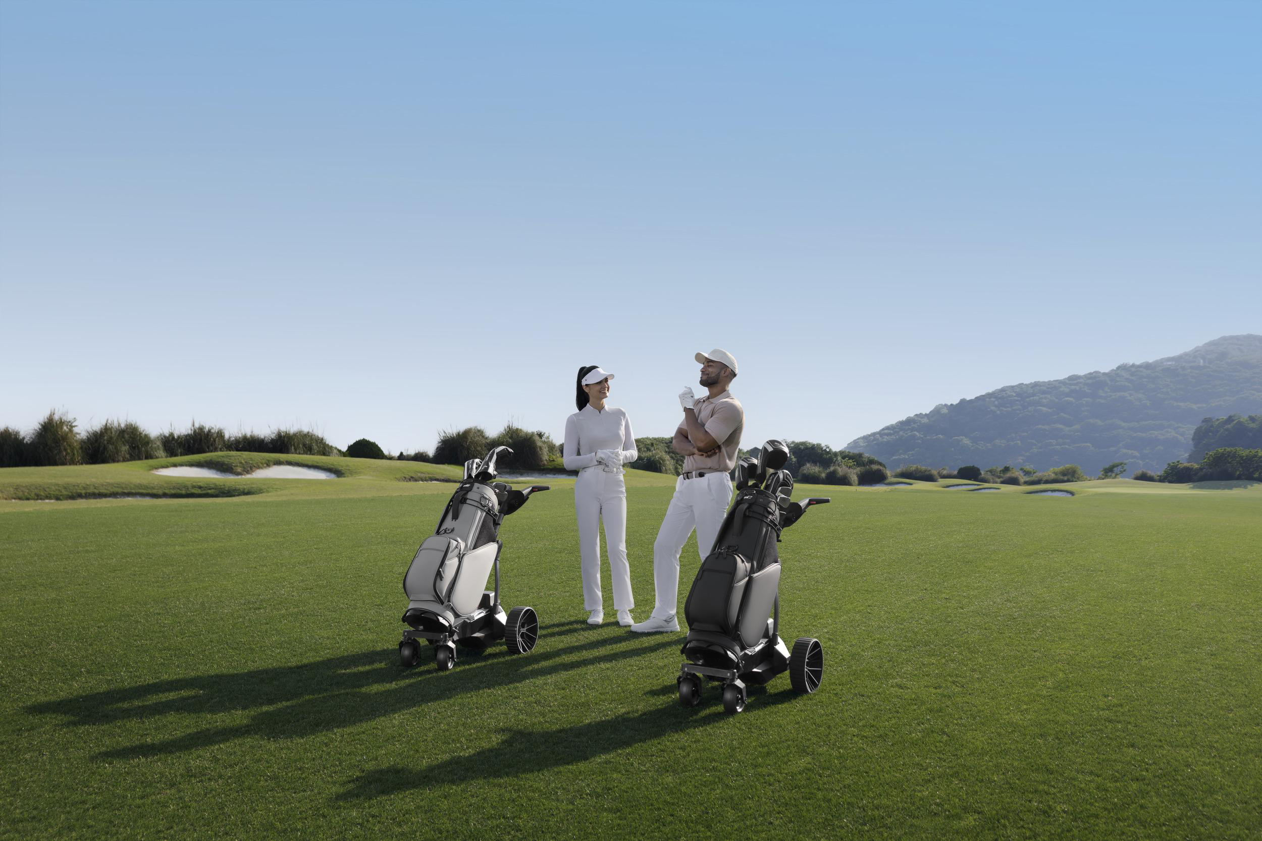 Zwei Golfer stehen auf einem grünen Golfplatz neben ihren Trolleys, im Hintergrund bewaldete Hügel und blauer Himmel.