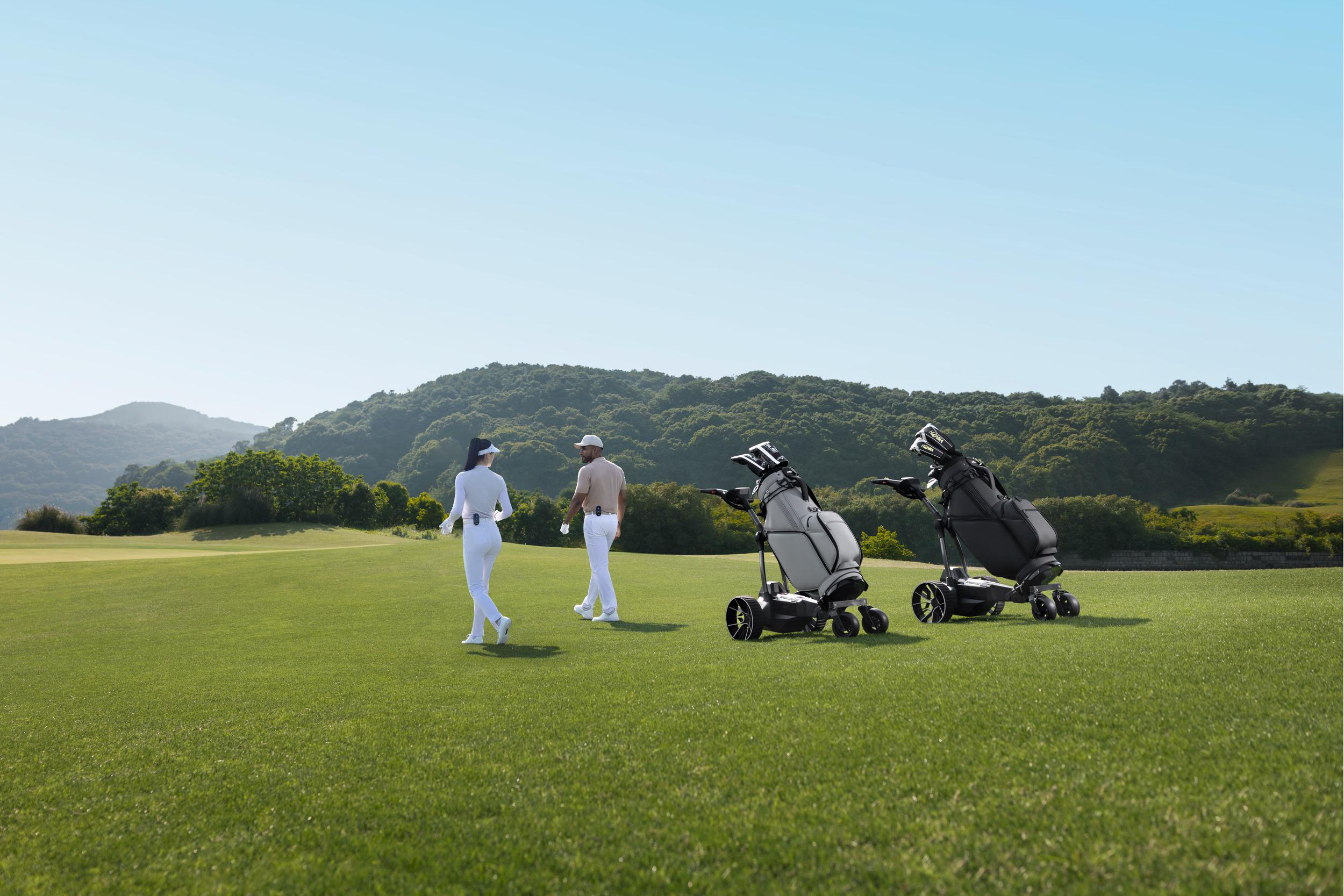 Zwei Golfer gehen mit zwei motorisierten Golfbags auf einem grünen Platz vor bewaldeten Hügeln spazieren.