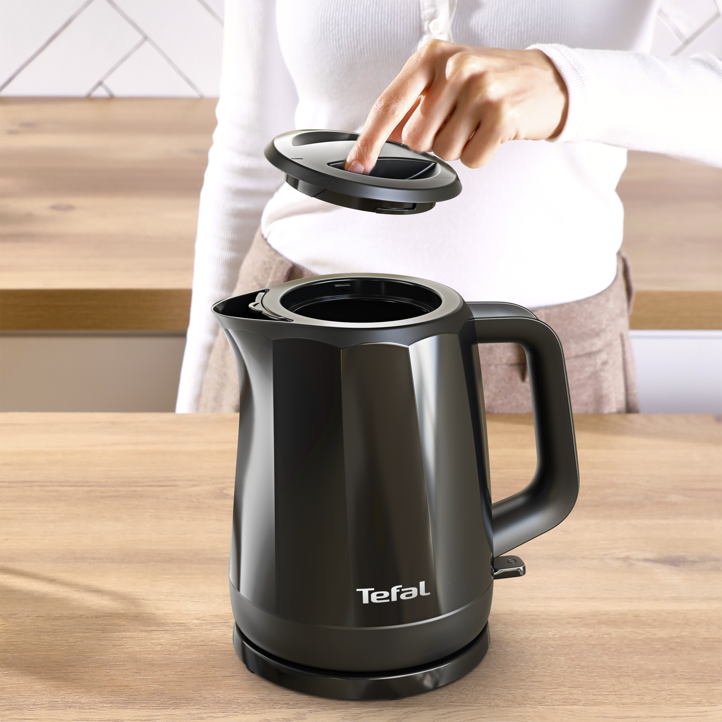 Egy személy kinyitja egy fekete Tefal vízforraló fedelét egy fa munkalapon.