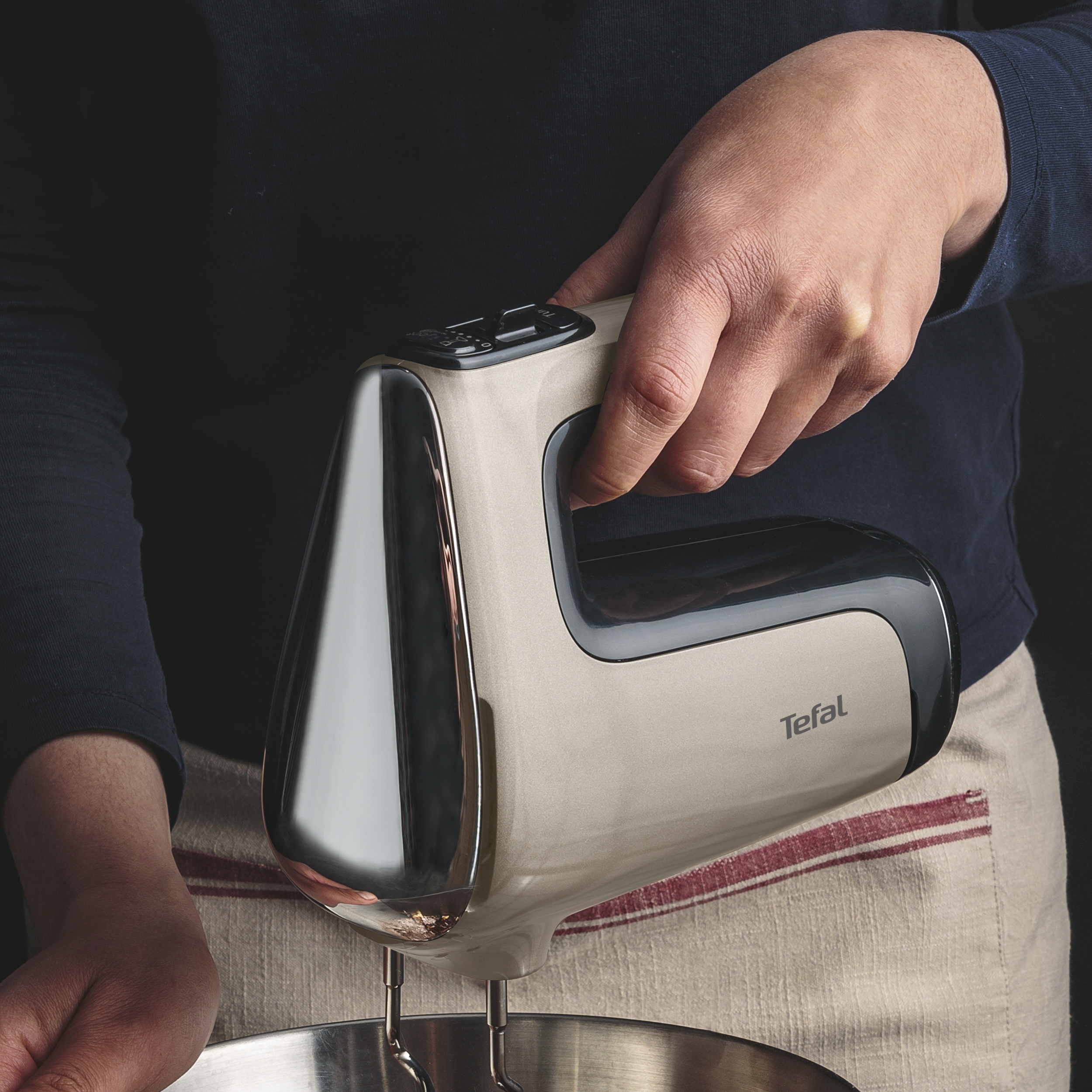 Egy személy bézs és fekete Tefal kézi mixert tart egy fém tál fölött.