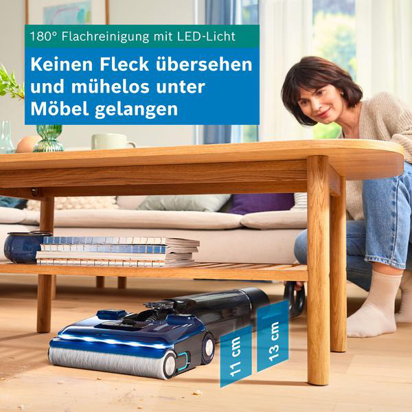 Ein blauer Staubsauger reinigt flach unter einem Holztisch, während eine Frau zuschaut.