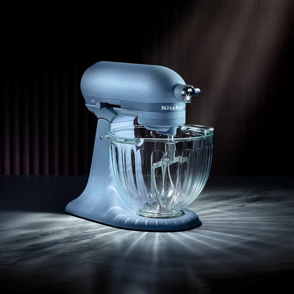 Ein blauer KitchenAid Standmixer mit einer Glasschüssel steht auf einer Marmorarbeitsplatte unter einem Lichtstrahl.
