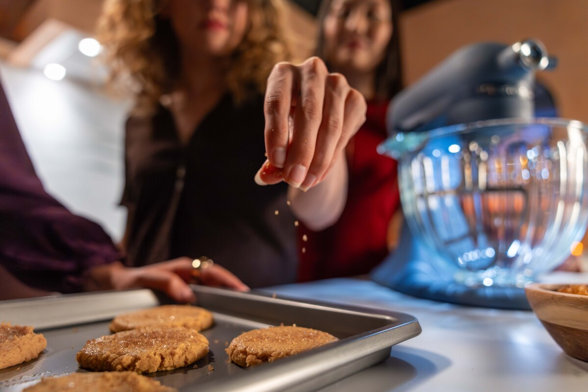 Eine Frau streut Gewürze auf frisch gebackene Plätzchen auf einem Blech, mit einem Mixer im Hintergrund.