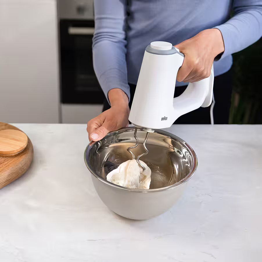 Una persona usa un mixer a mano bianco per mescolare l'impasto in una ciotola d'acciaio.