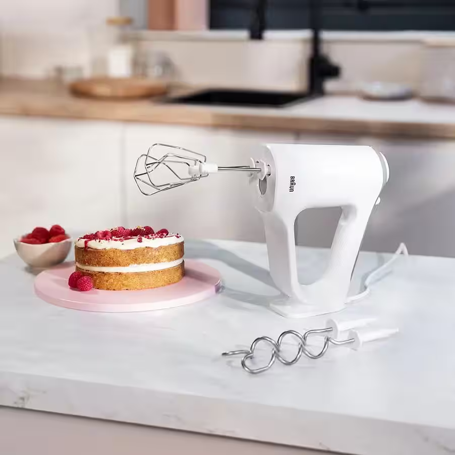 Un mixer a mano bianco con fruste poggia su un bancone accanto a una torta a strati e ganci.