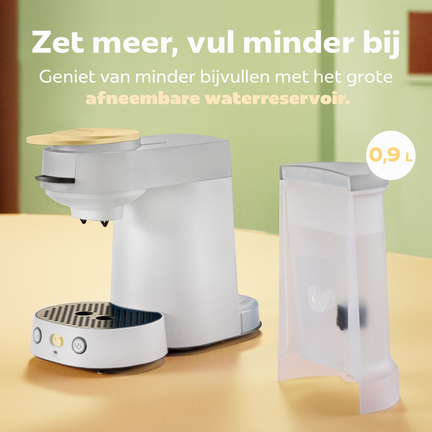 Een witte koffiemachine met een afneembaar waterreservoir van 0,9 liter op een tafel tegen een groene achtergrond.