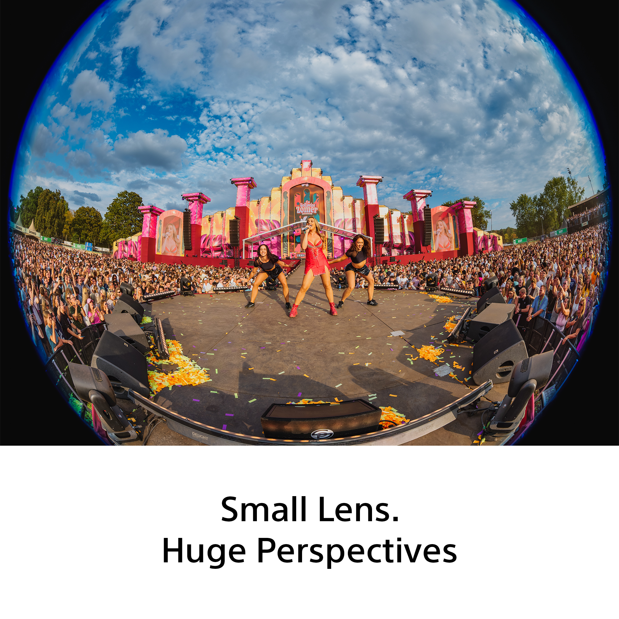 Vue fisheye d'un concert avec trois artistes sur scène, une foule nombreuse, un décor coloré et un ciel bleu.