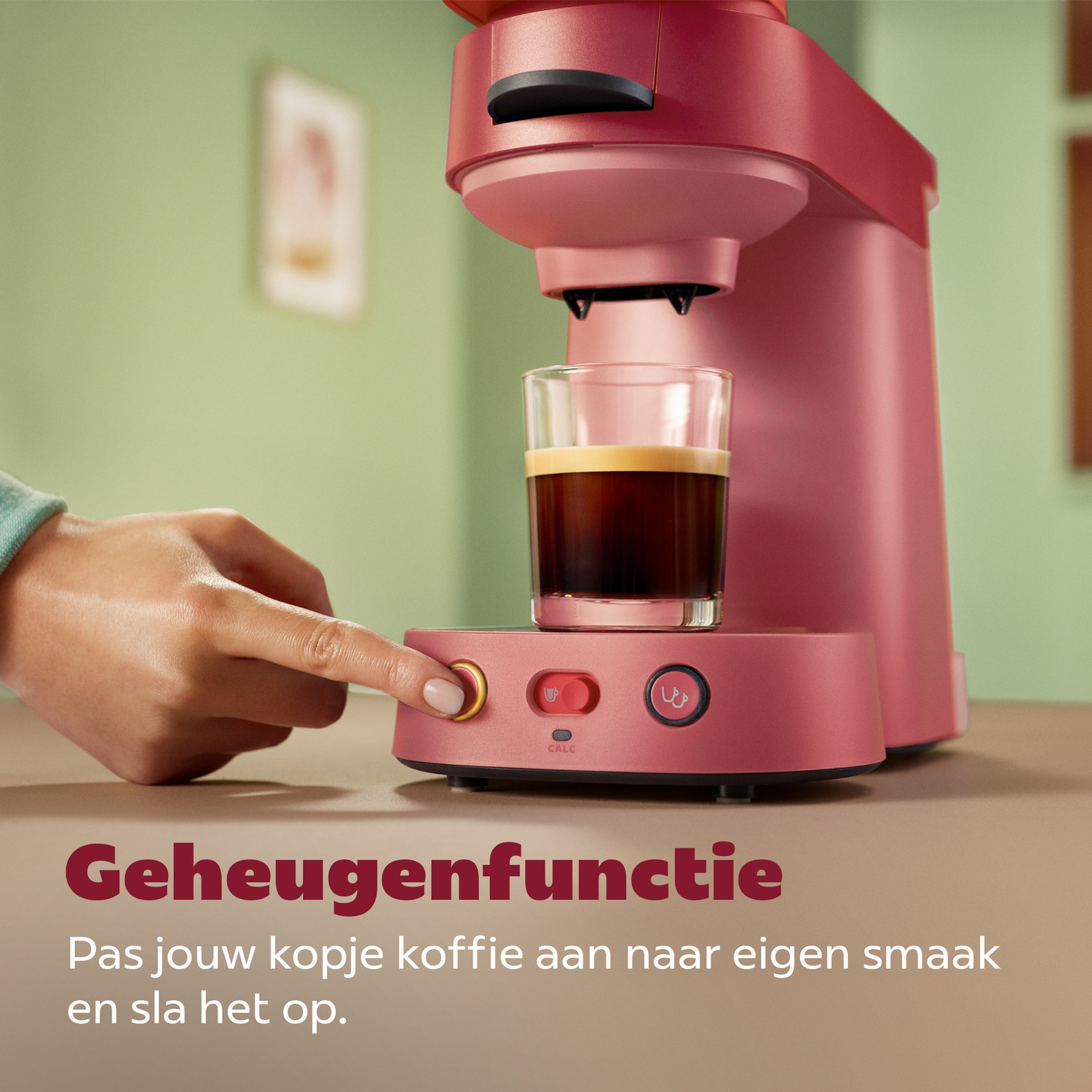 Een hand drukt op de knop van een roze koffiezetapparaat met een kop koffie.