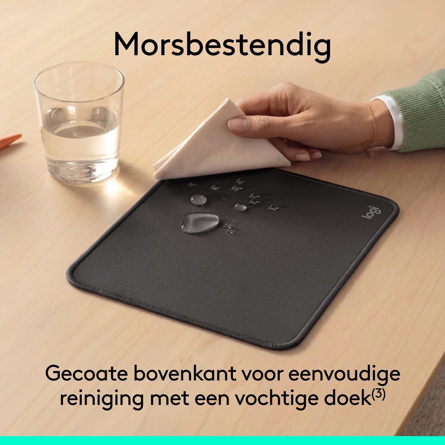 Une main essuie des gouttes d'eau d'un tapis de souris noir avec un chiffon, à côté d'un verre d'eau sur une table en bois.