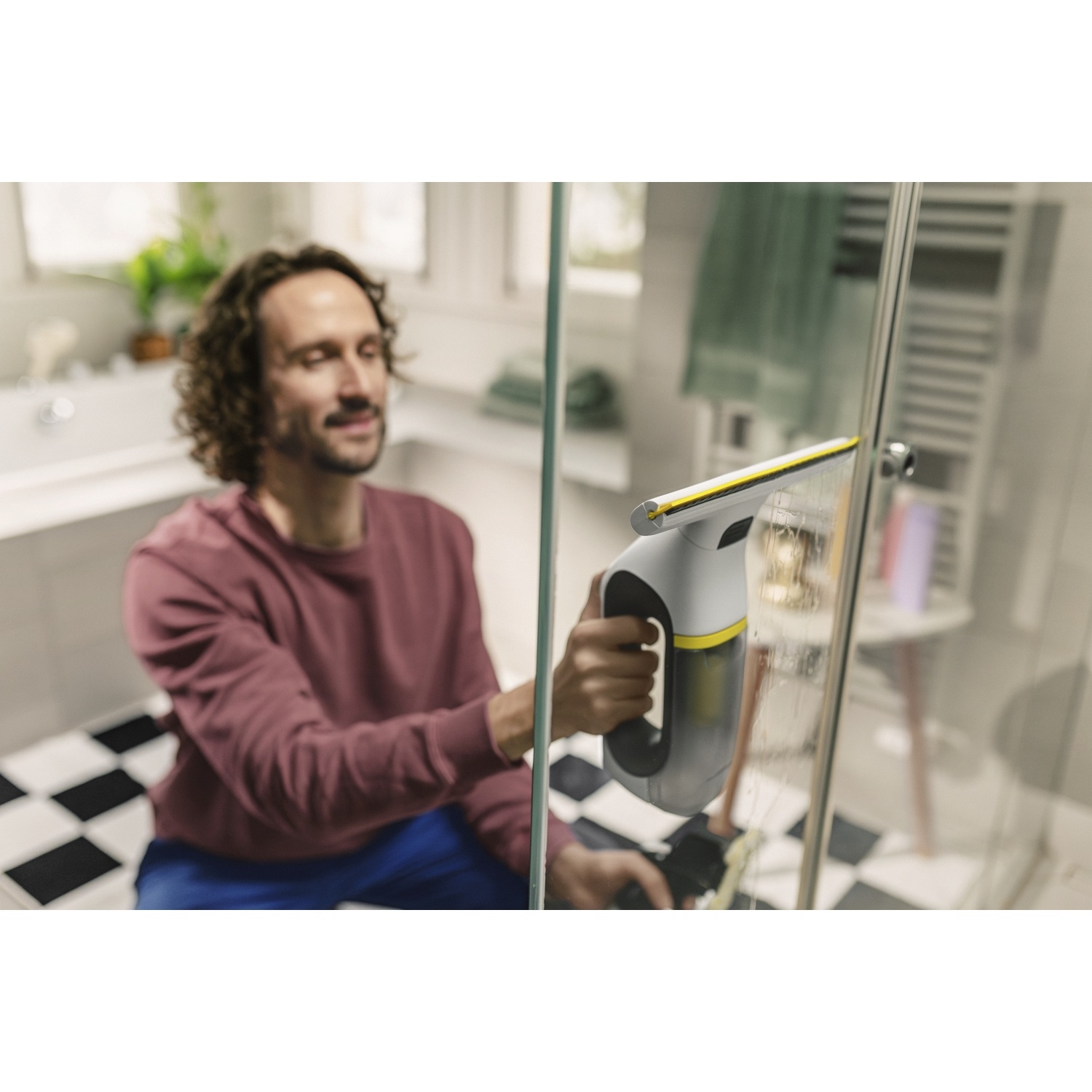 Un homme aux longs cheveux bouclés nettoie une porte de douche en verre avec un nettoyeur de vitres.