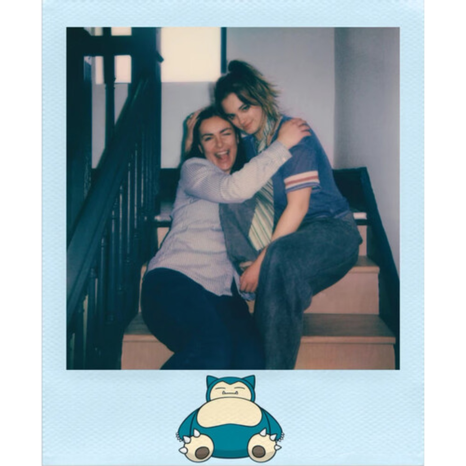 Two smiling young women embrace while sitting on indoor stairs next to a dark railing, with a Snorlax illustration below.