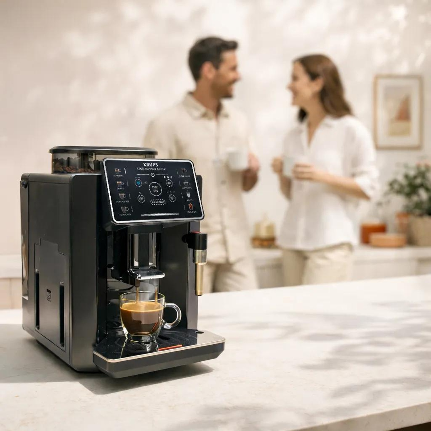 Une machine à café Krups noire sur un comptoir de cuisine, avec un couple souriant en arrière-plan flou.