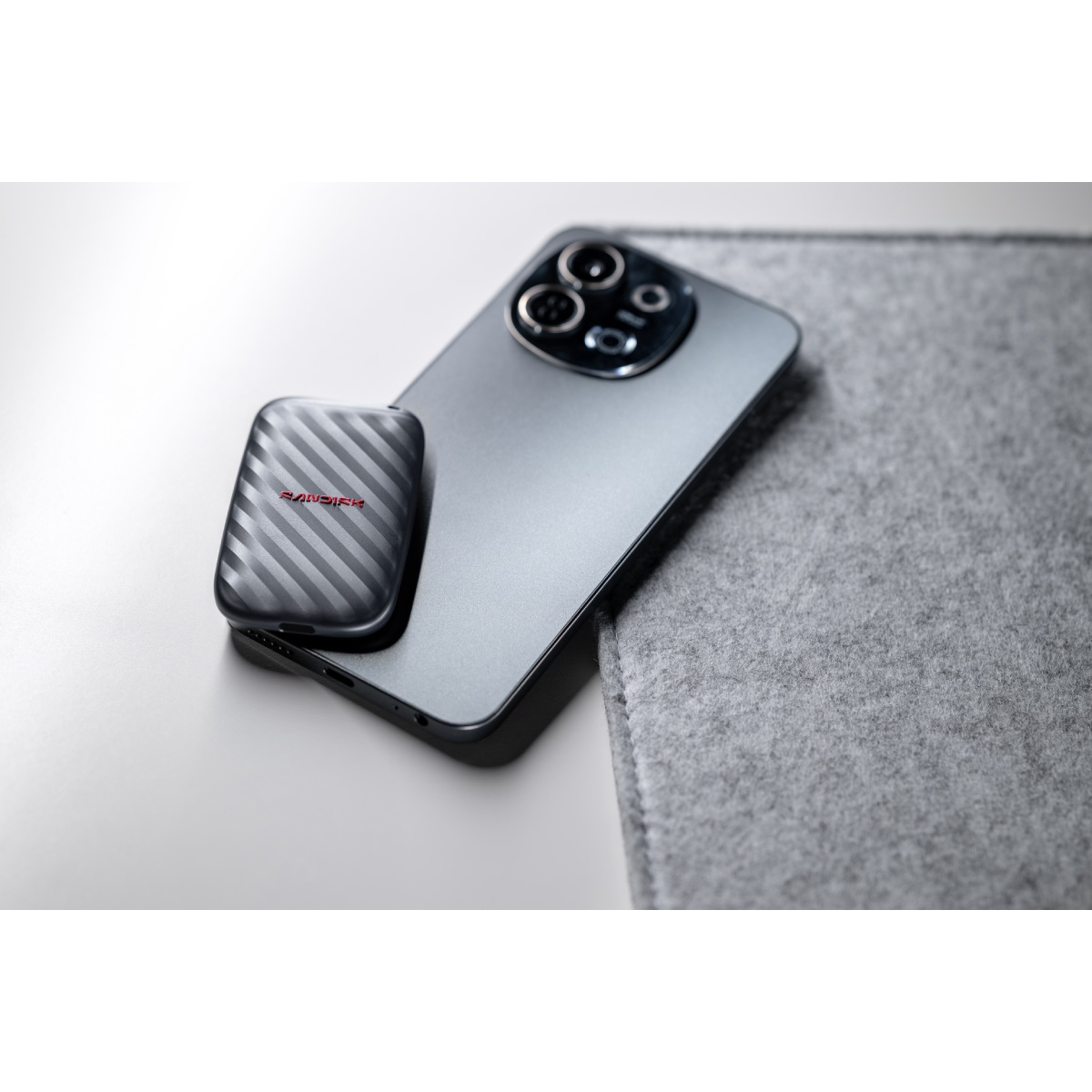 Un SSD portátil SanDisk oscuro reposa sobre un teléfono inteligente moderno junto a una alfombrilla de fieltro gris.