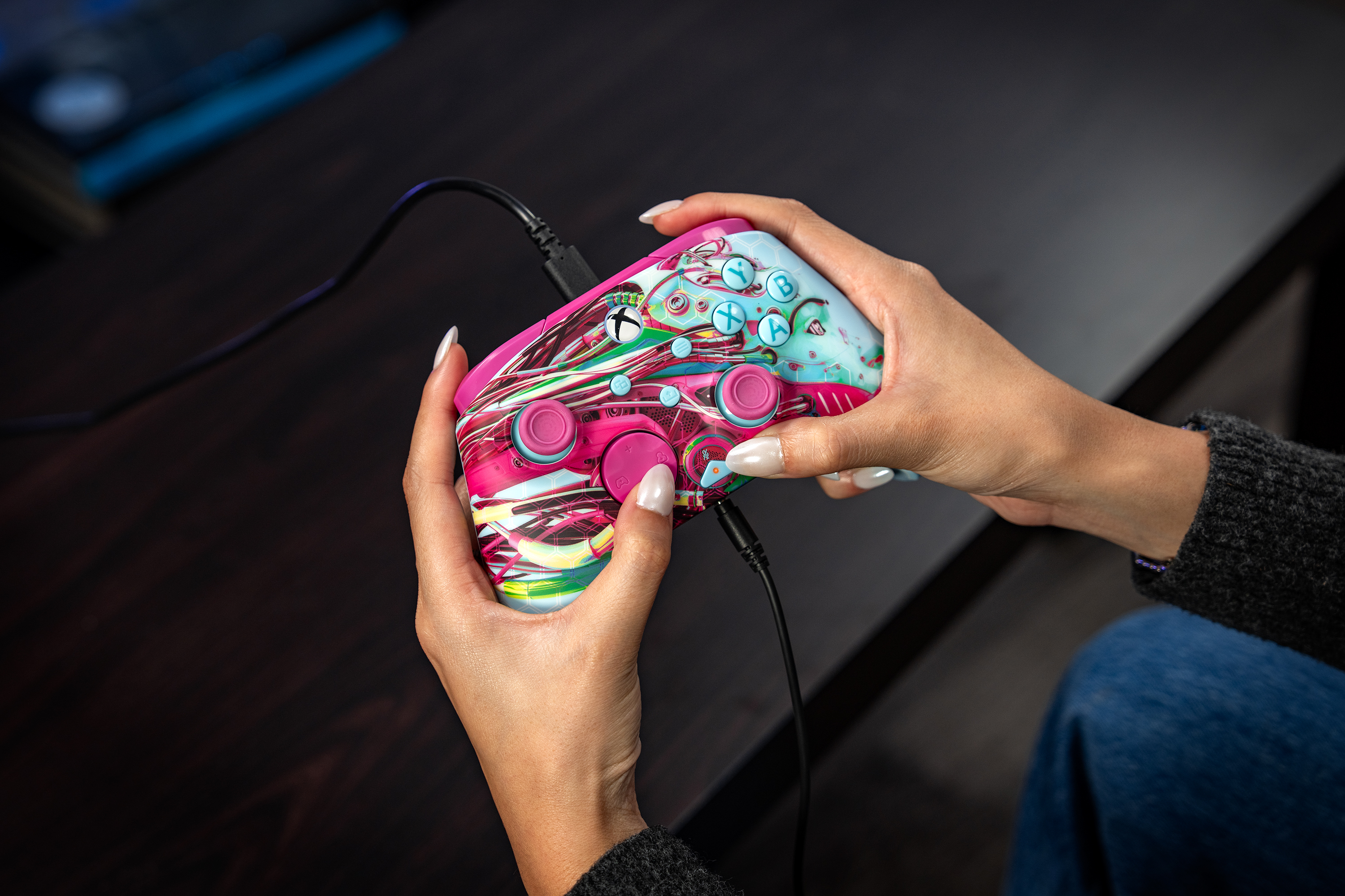Mains tenant une manette Xbox rose et colorée avec fil sur une table.