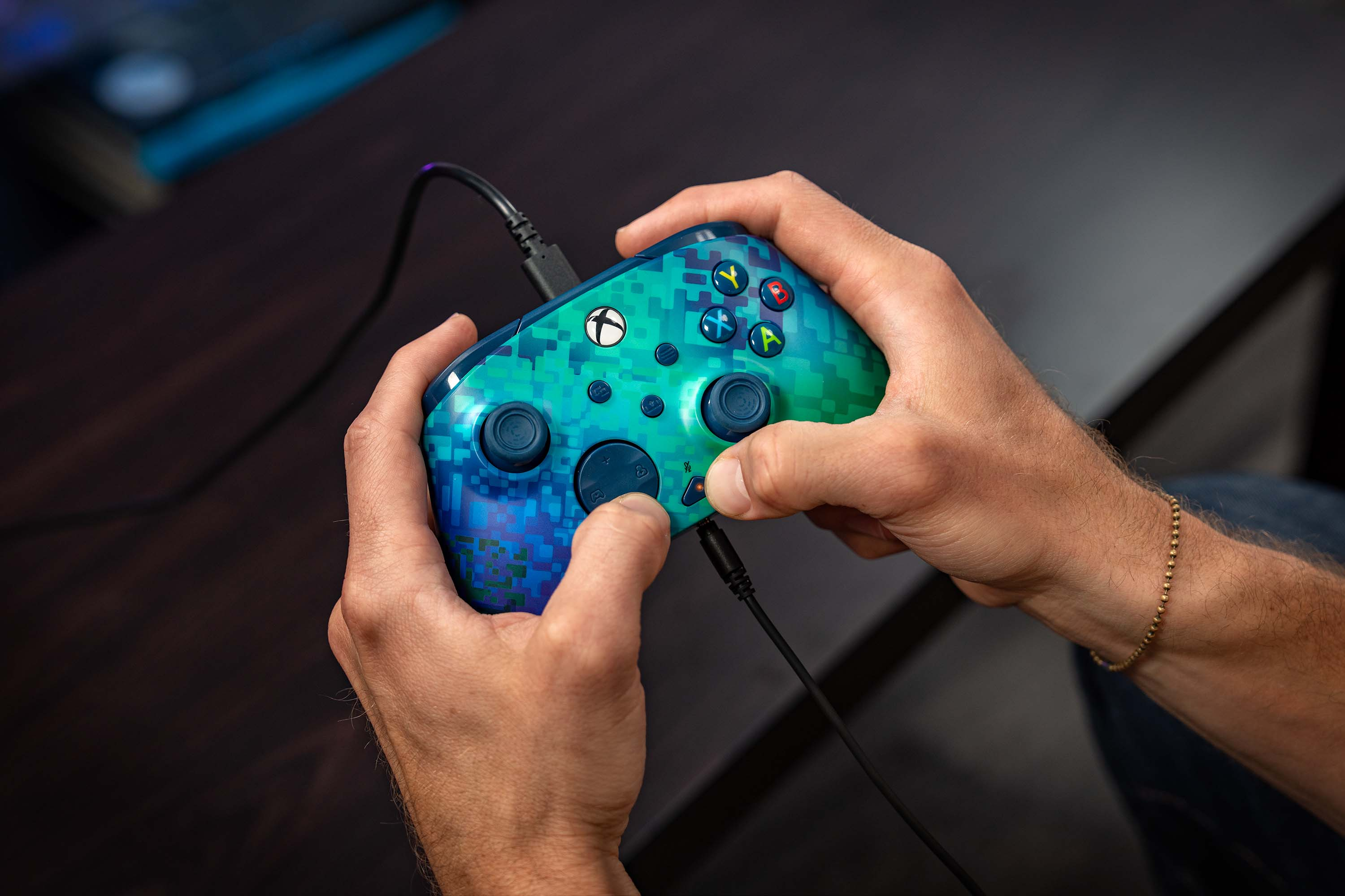 Personne tenant une manette Xbox pixelisée bleue et verte avec un câble de charge branché.