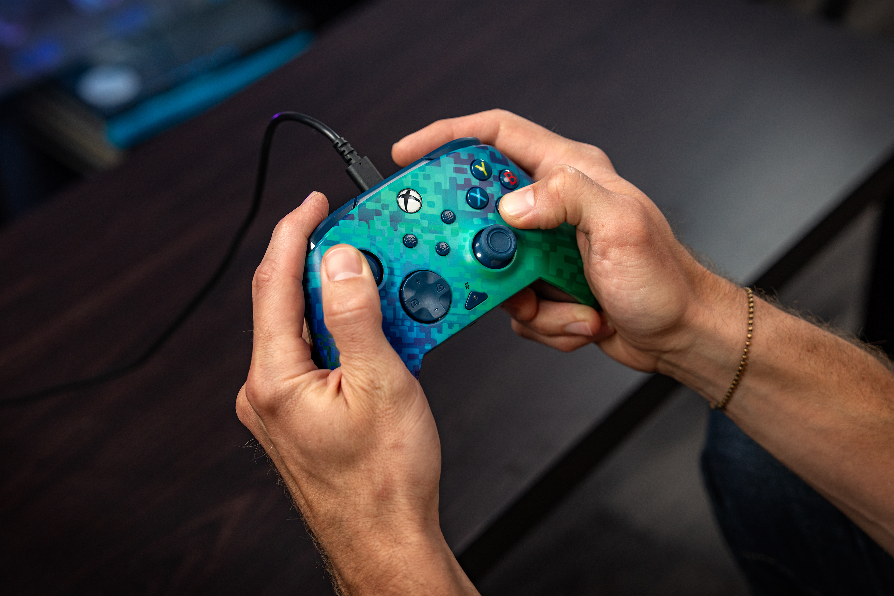 Une personne tient une manette Xbox pixélisée bleue et verte sur un bureau en bois sombre.