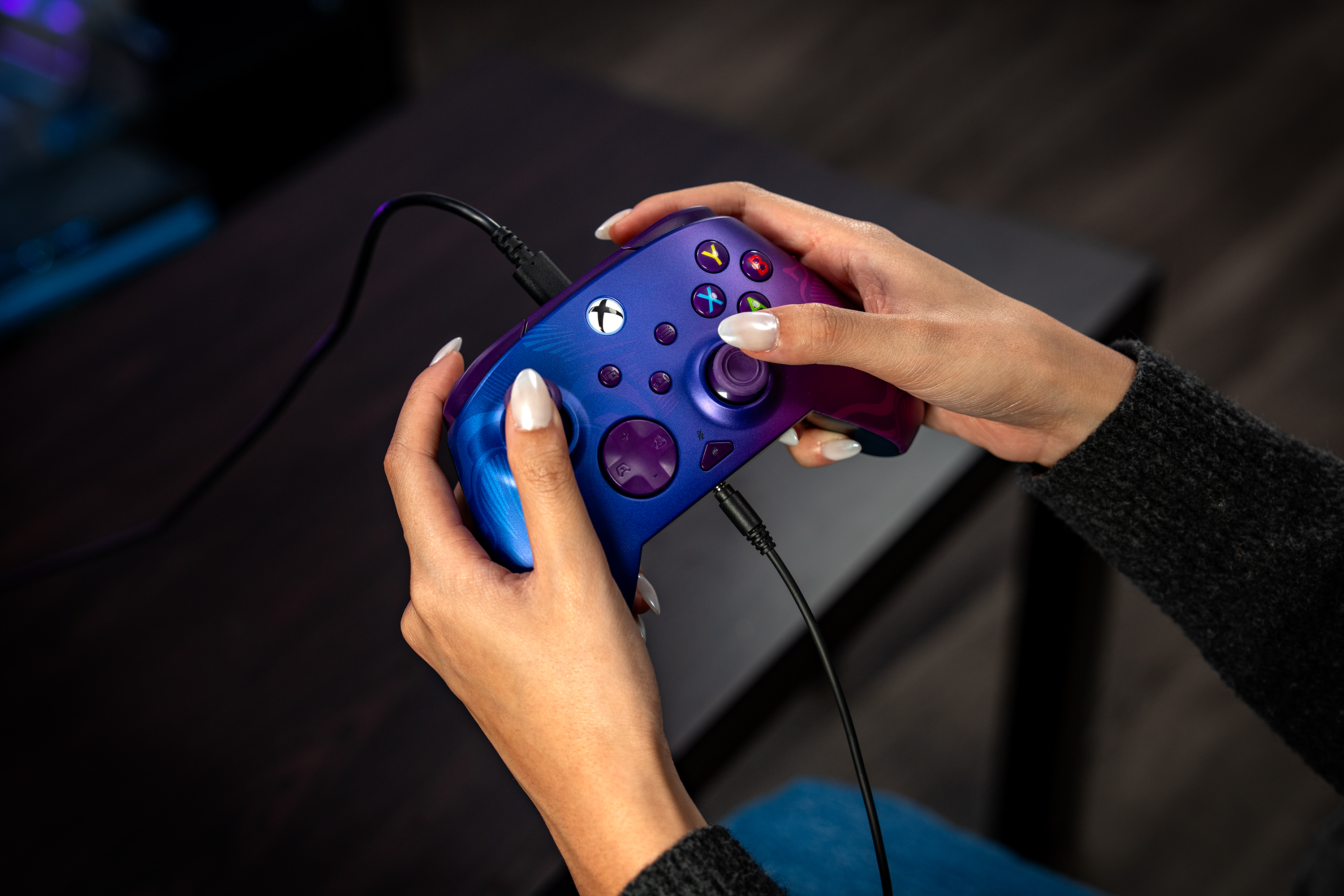 Personne tenant une manette Xbox bleue et violette avec un cordon attaché assise à une table.
