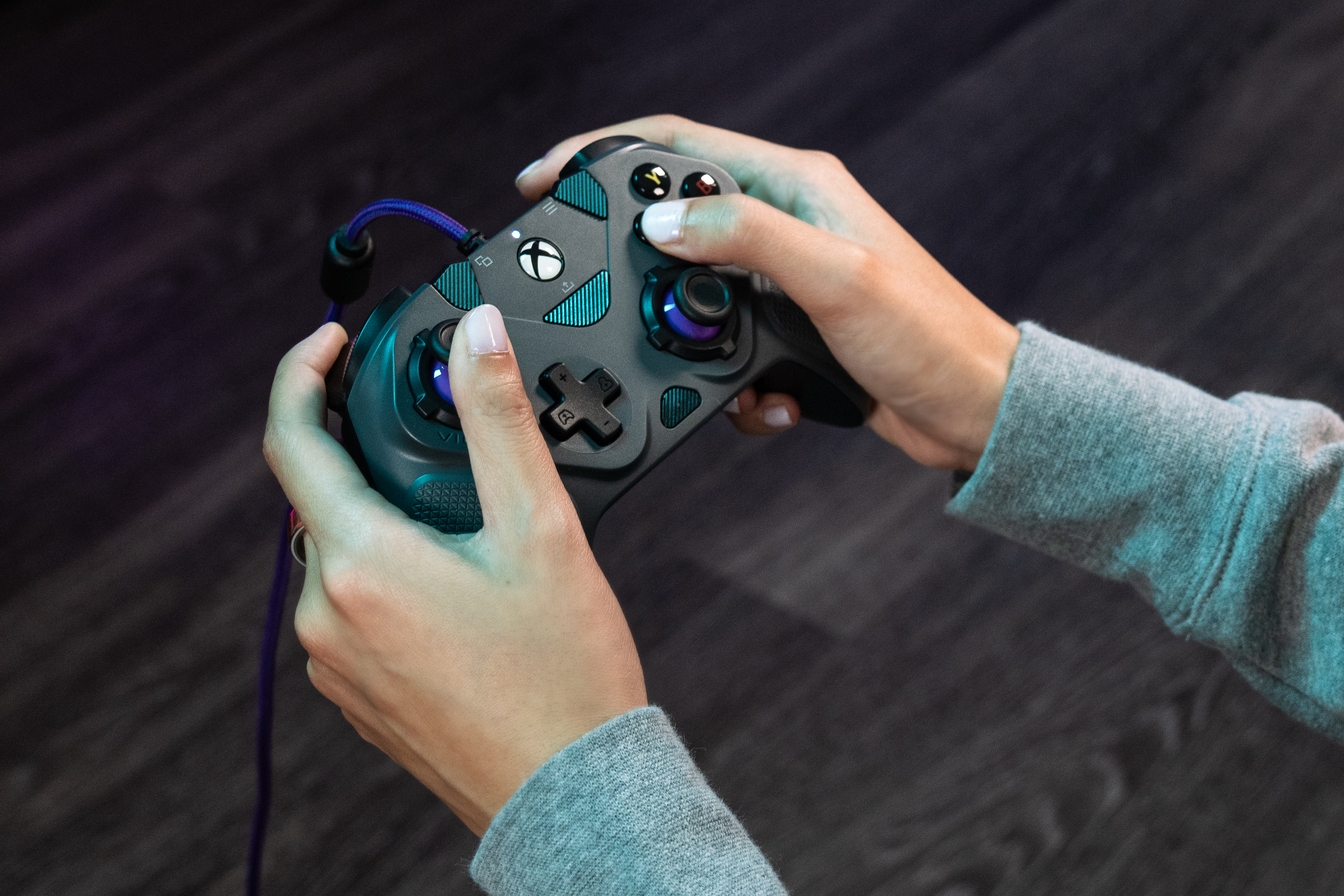 Personne en pull gris tenant une manette Xbox noire sur fond de bois sombre.