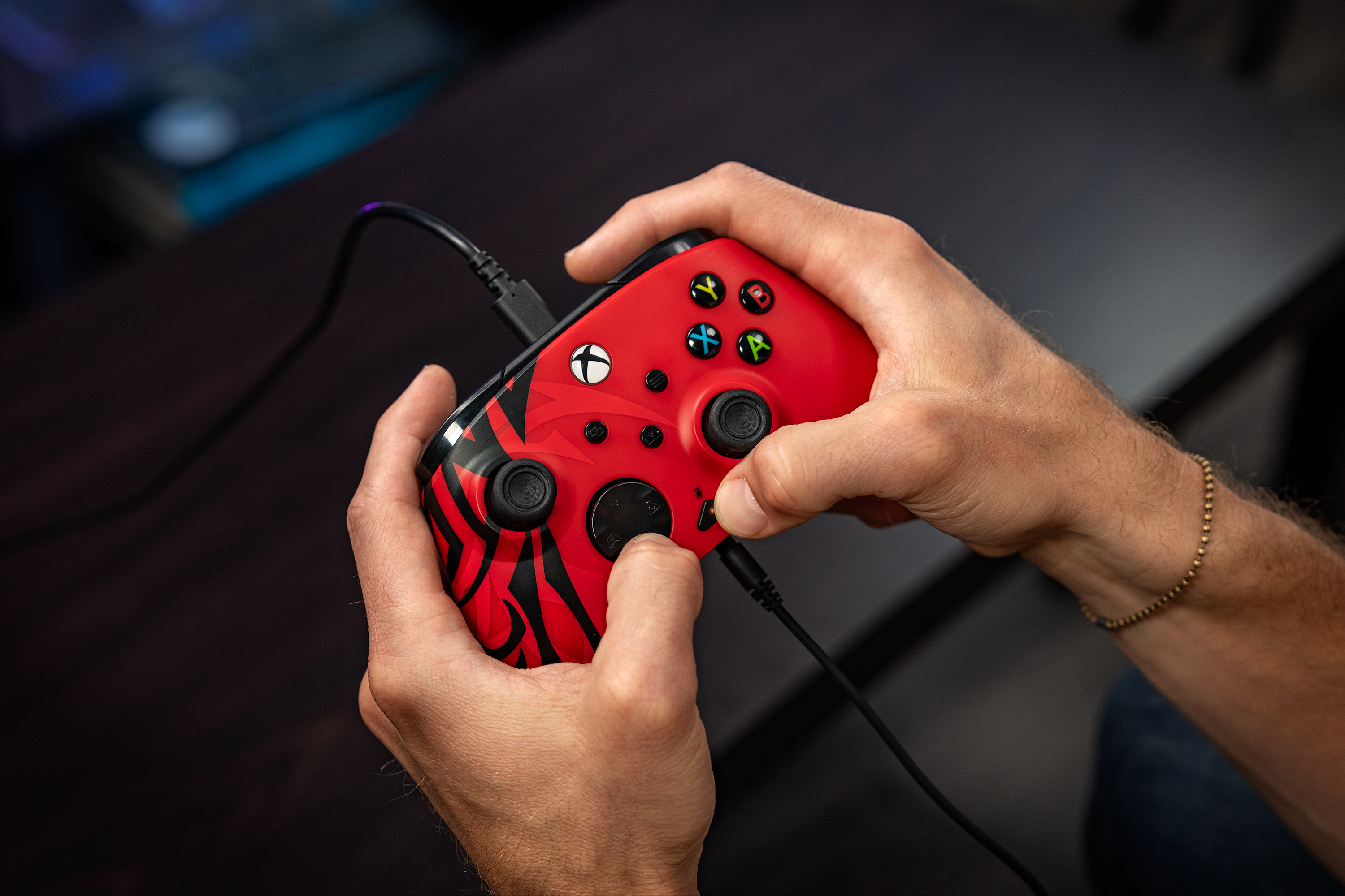 Personne tenant une manette Xbox rouge et noire connectée par un câble noir sur une table sombre.