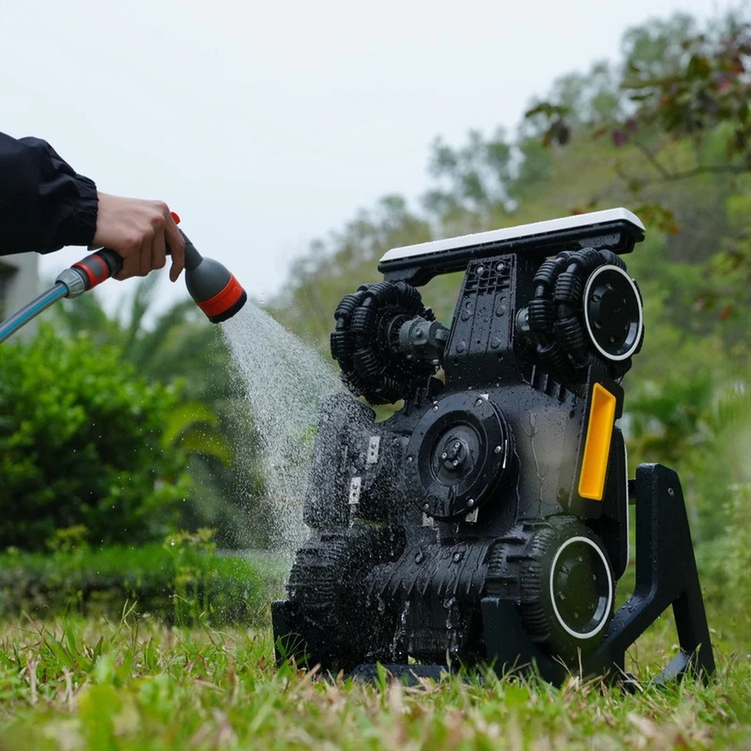 Qualcuno lava un robot tagliaerba nero sul prato con l'acqua di un tubo.