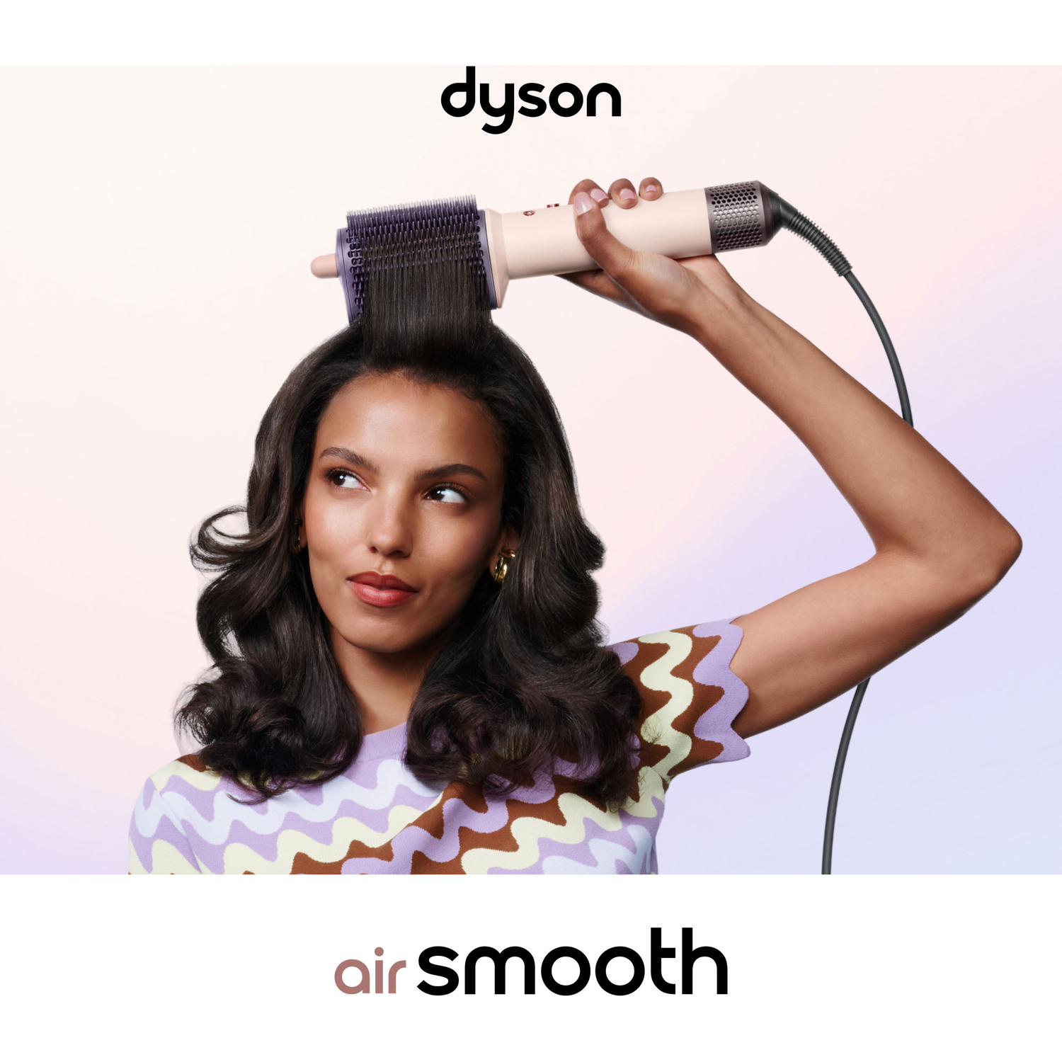 Een vrouw houdt een Dyson haarstyler vast en stijlt haar lange, donkere haar tegen een pastelkleurige achtergrond.