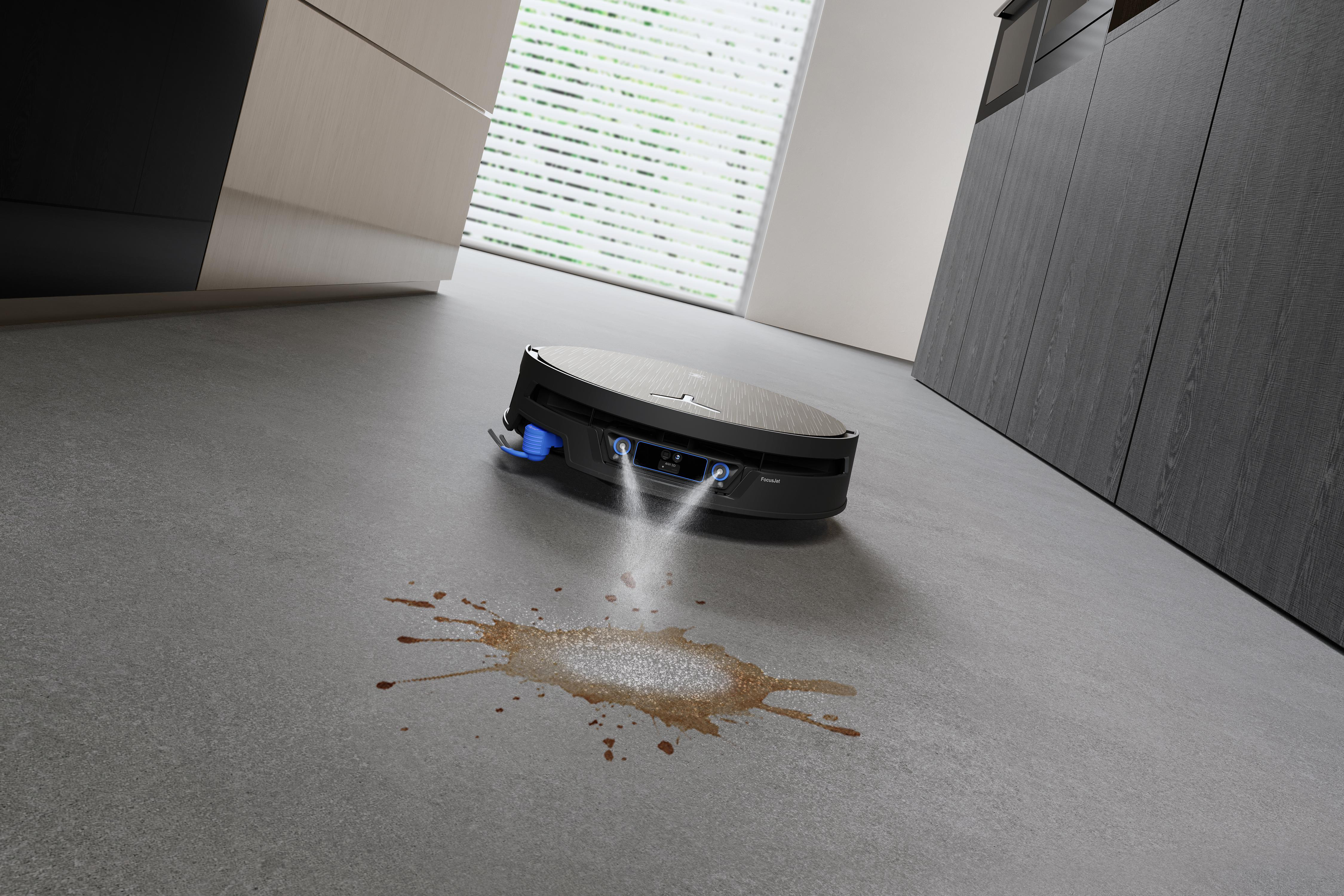 Ein schwarzer Saugroboter sprüht Wasser auf einen Kaffeefleck auf dem grauen Boden.