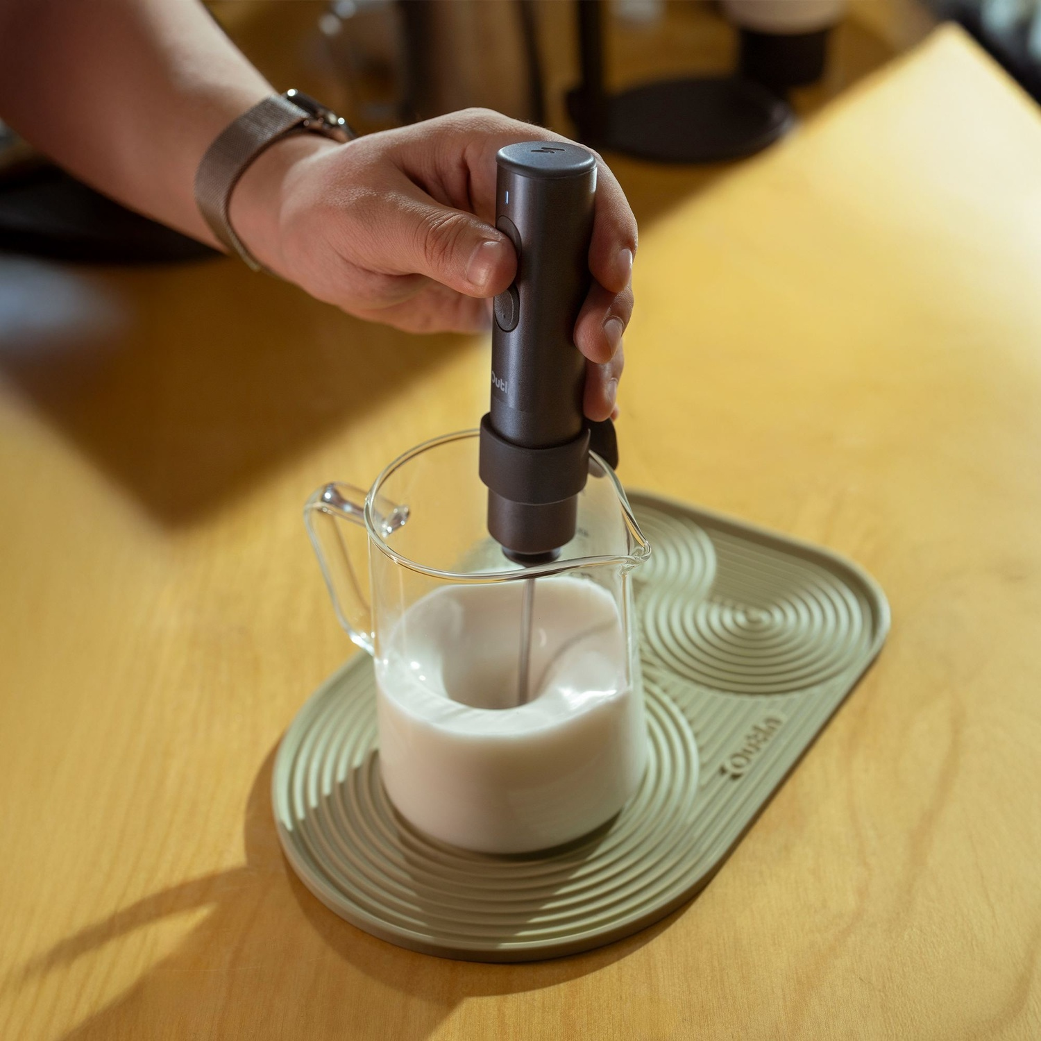 Una mano tiene un montalatte sopra una caraffa di vetro con latte su un tappetino.