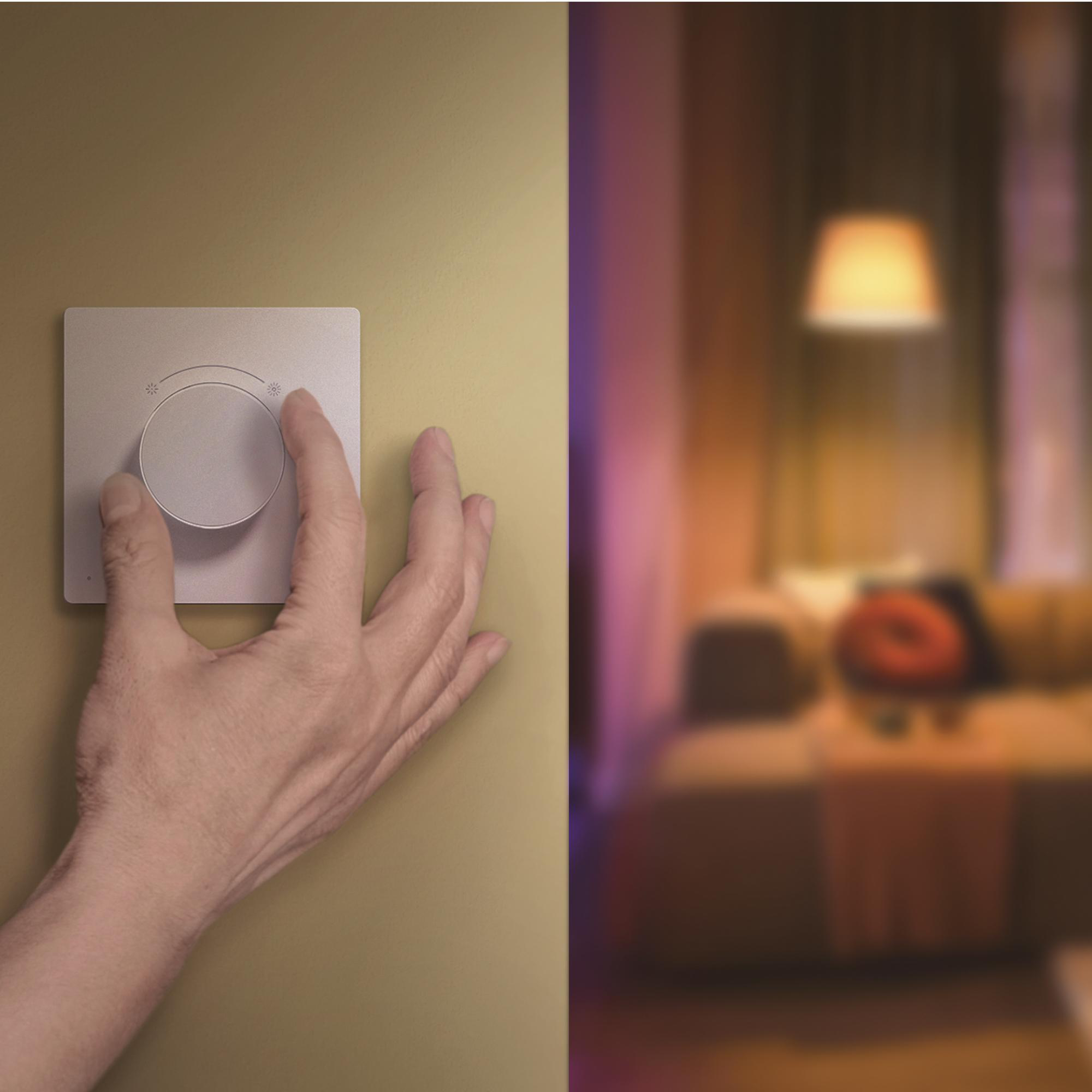 Eine Hand bedient einen runden Wandschalter vor einem verschwommenen Wohnzimmer.