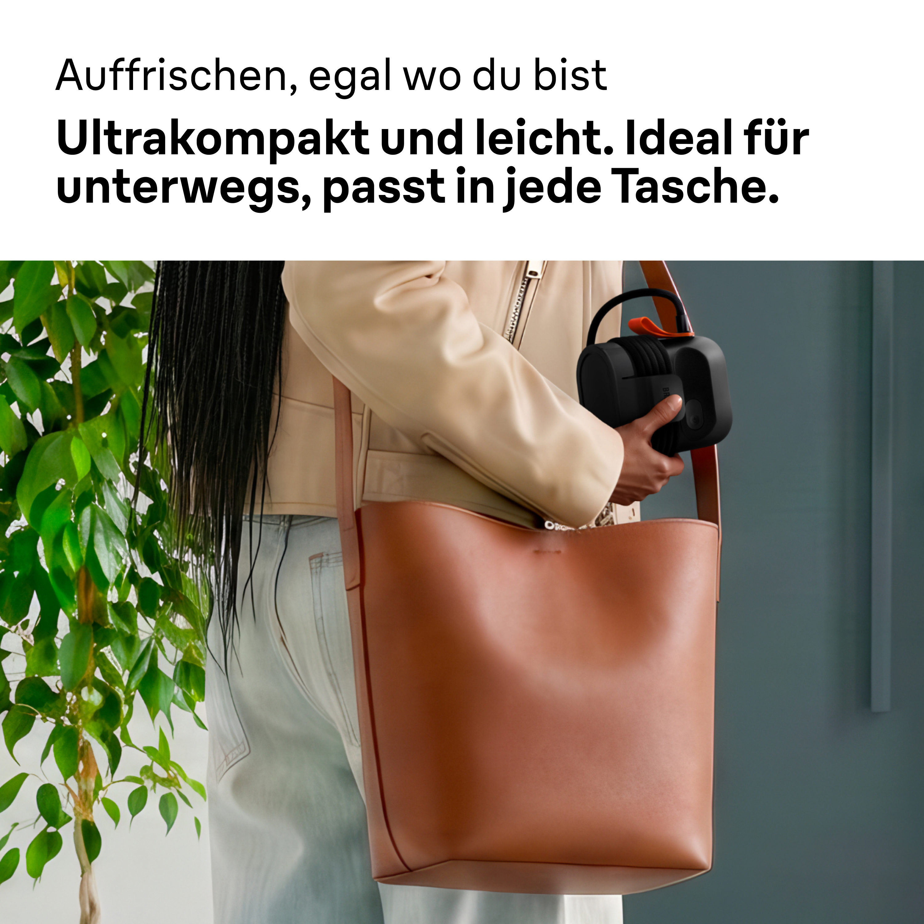 Eine Person mit Beiger Jacke hält ein kleines schwarzes Gerät neben einer großen braunen Tasche.