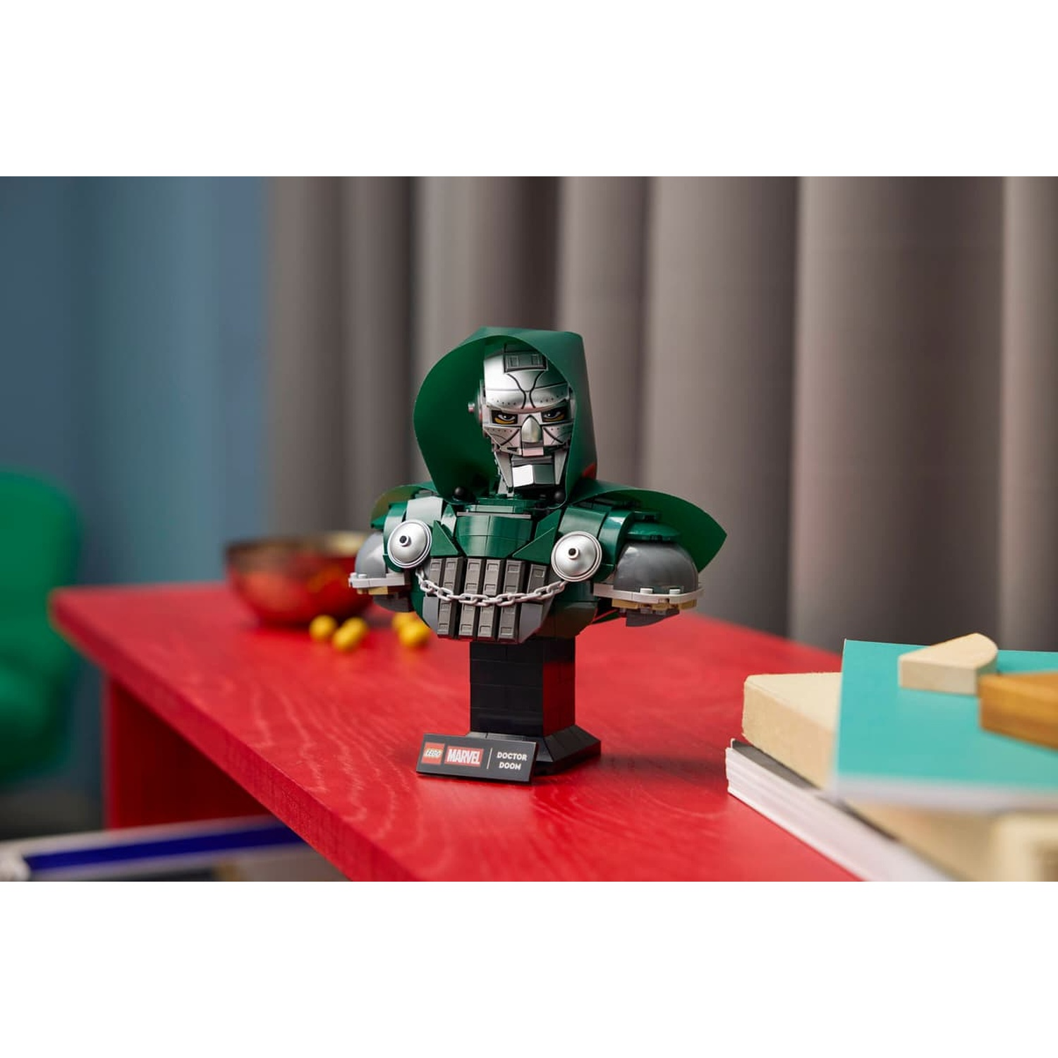Un buste Lego Marvel Doctor Doom repose sur une table rouge avec des livres et un bol en arrière-plan.