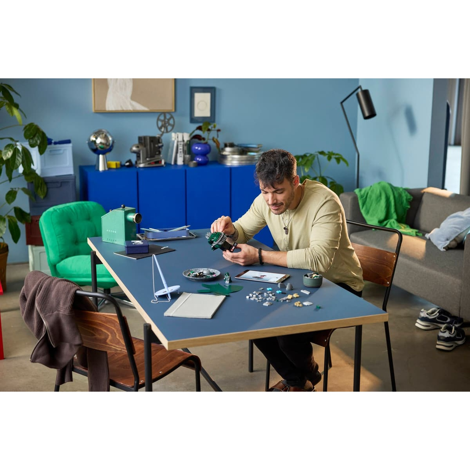 Un homme est assis à une table bleue dans un salon, assemblant un modèle Lego.