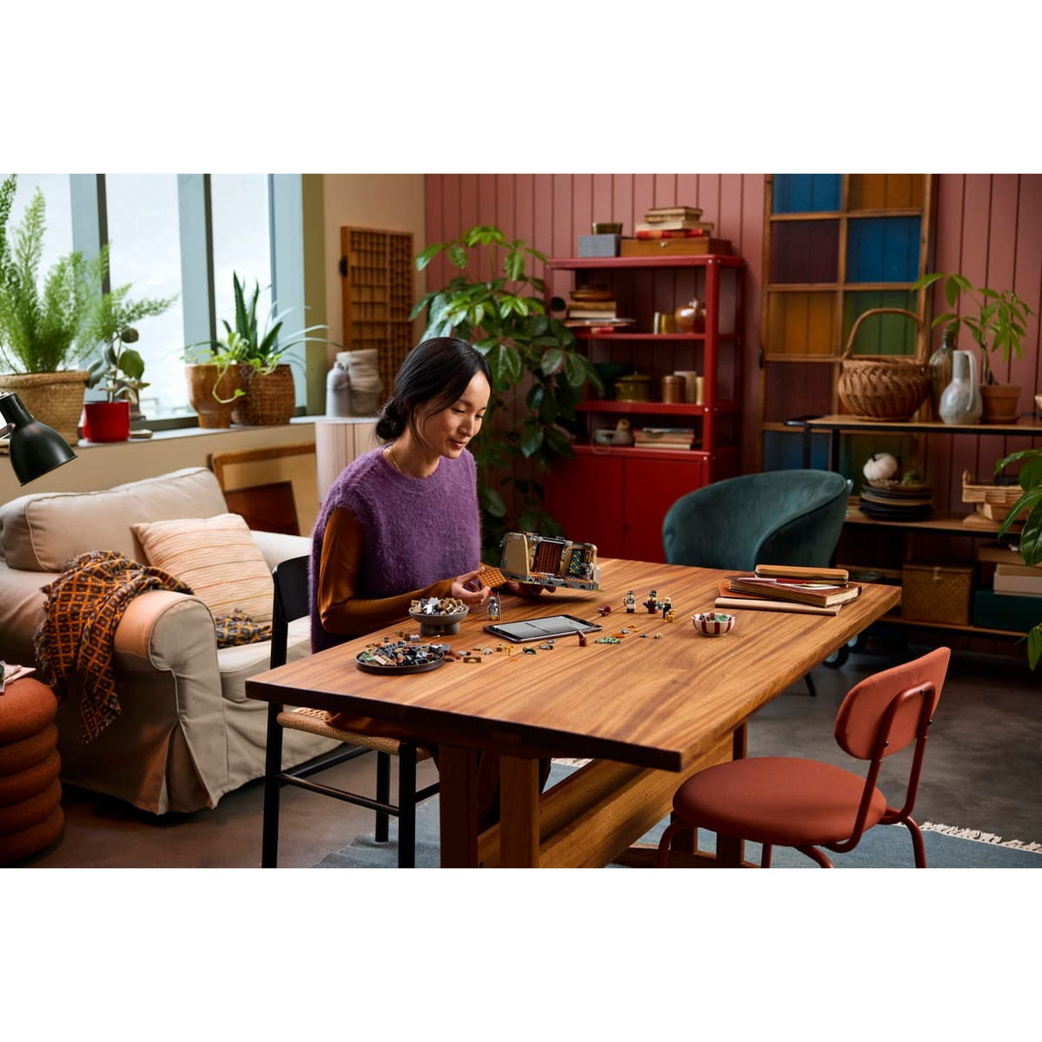 A woman sits at a wooden table in a living room, building a Lego set.