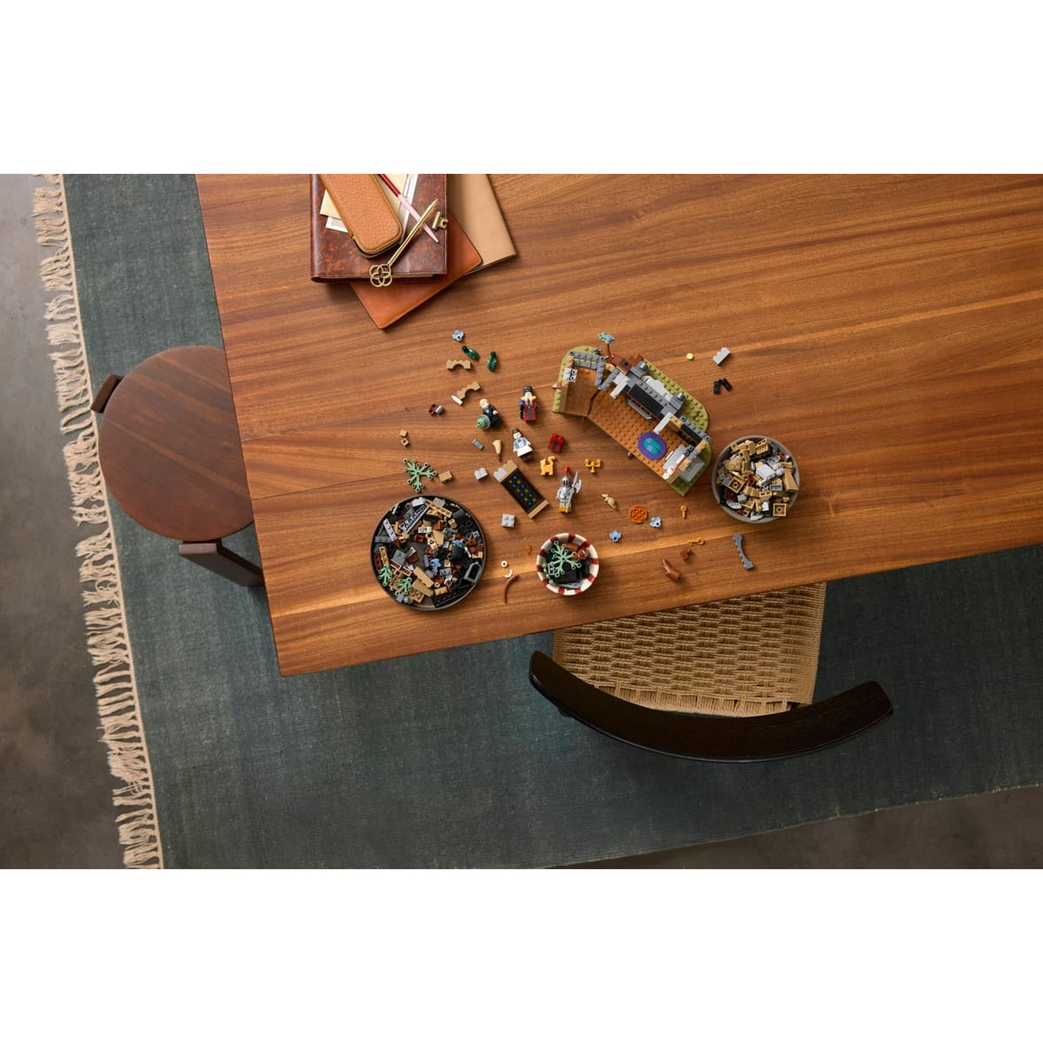 Overhead view of a wooden table with a Lego set, minifigures, bowls of bricks, a notebook, and chairs.
