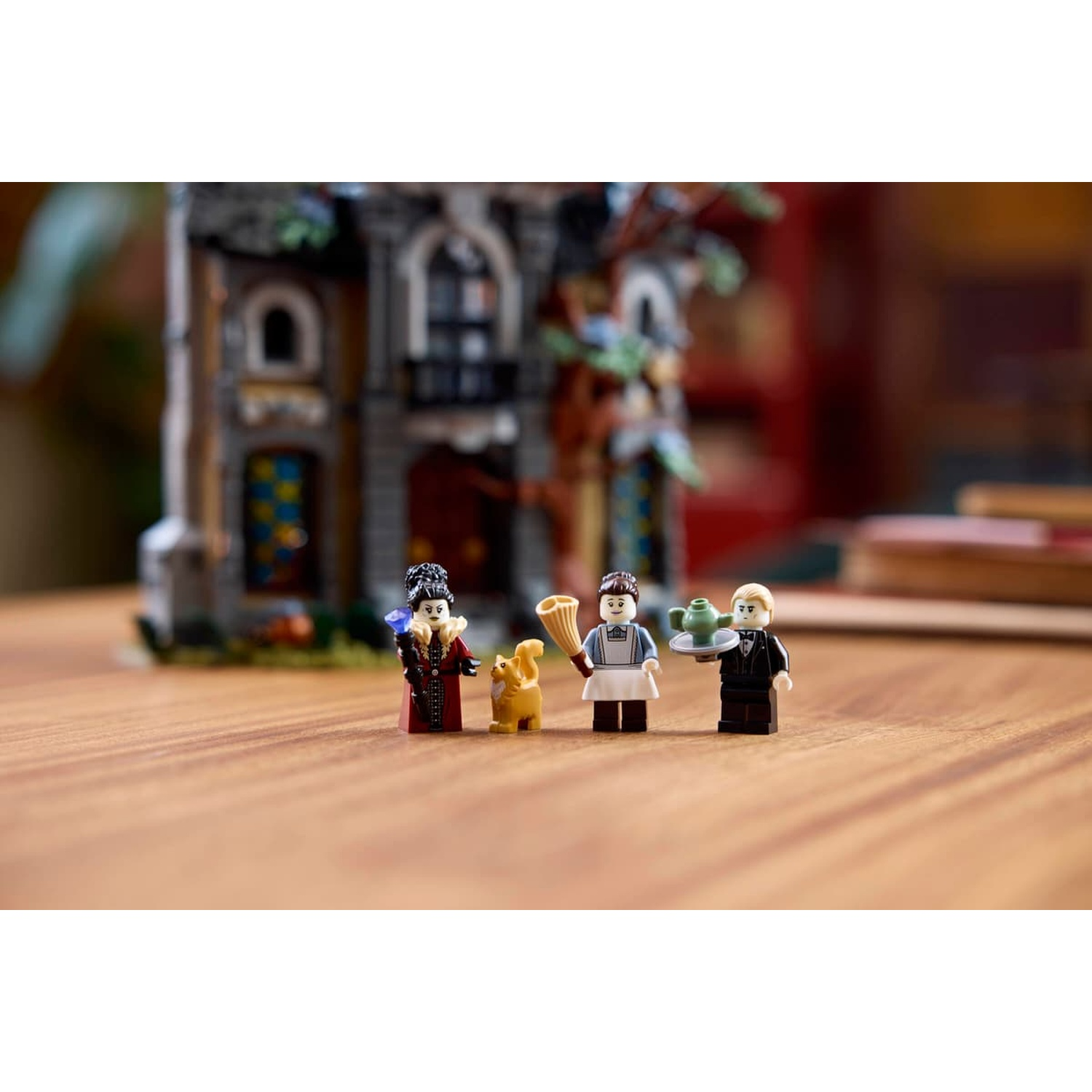 Three LEGO minifigures and a cat stand in front of a castle-like building on a wooden table.
