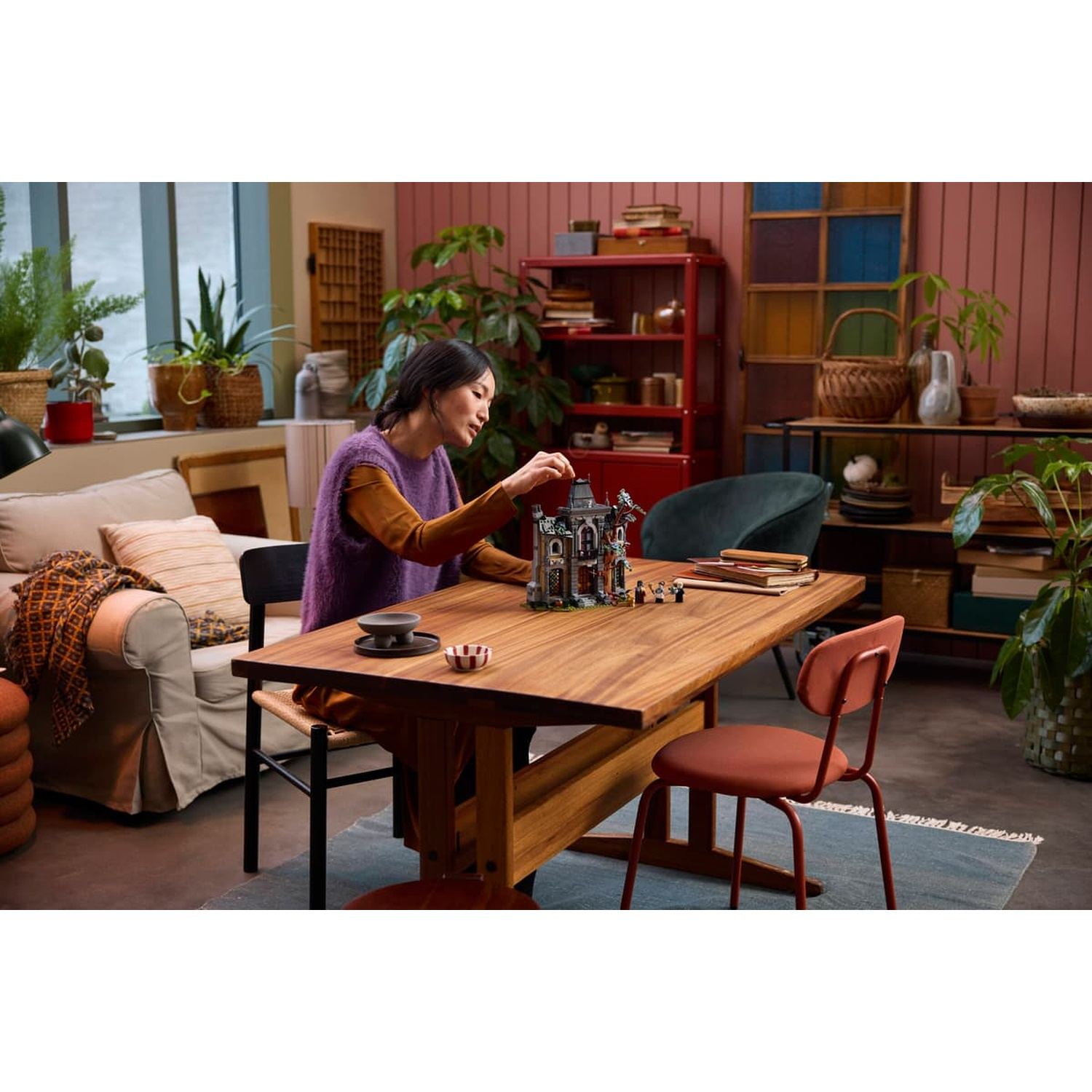 A woman sits at a wooden table in a cozy room, assembling a detailed LEGO model.
