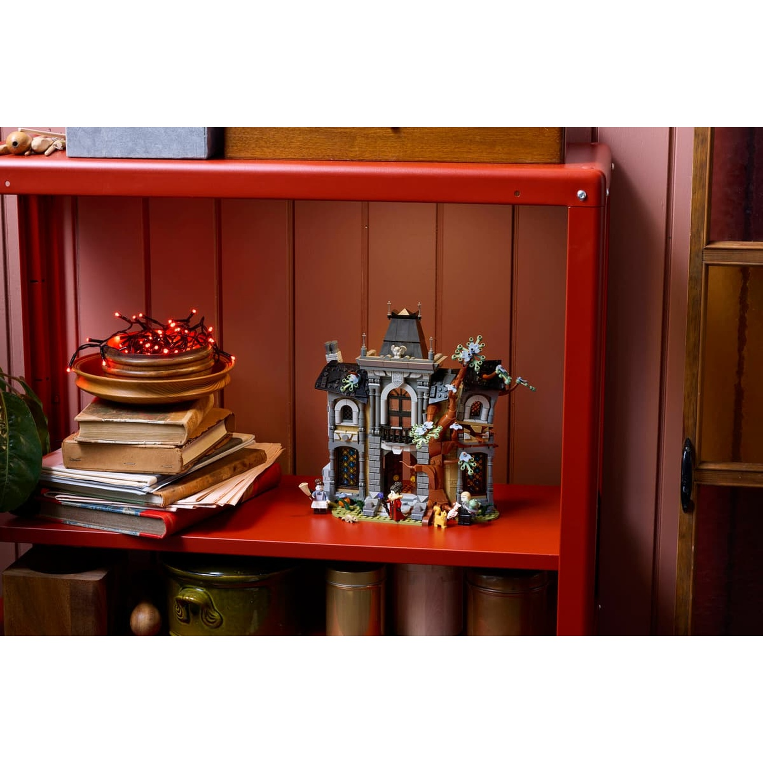 A Lego haunted mansion with mini figures on a red shelf, next to stacked books and glowing red fairy lights.