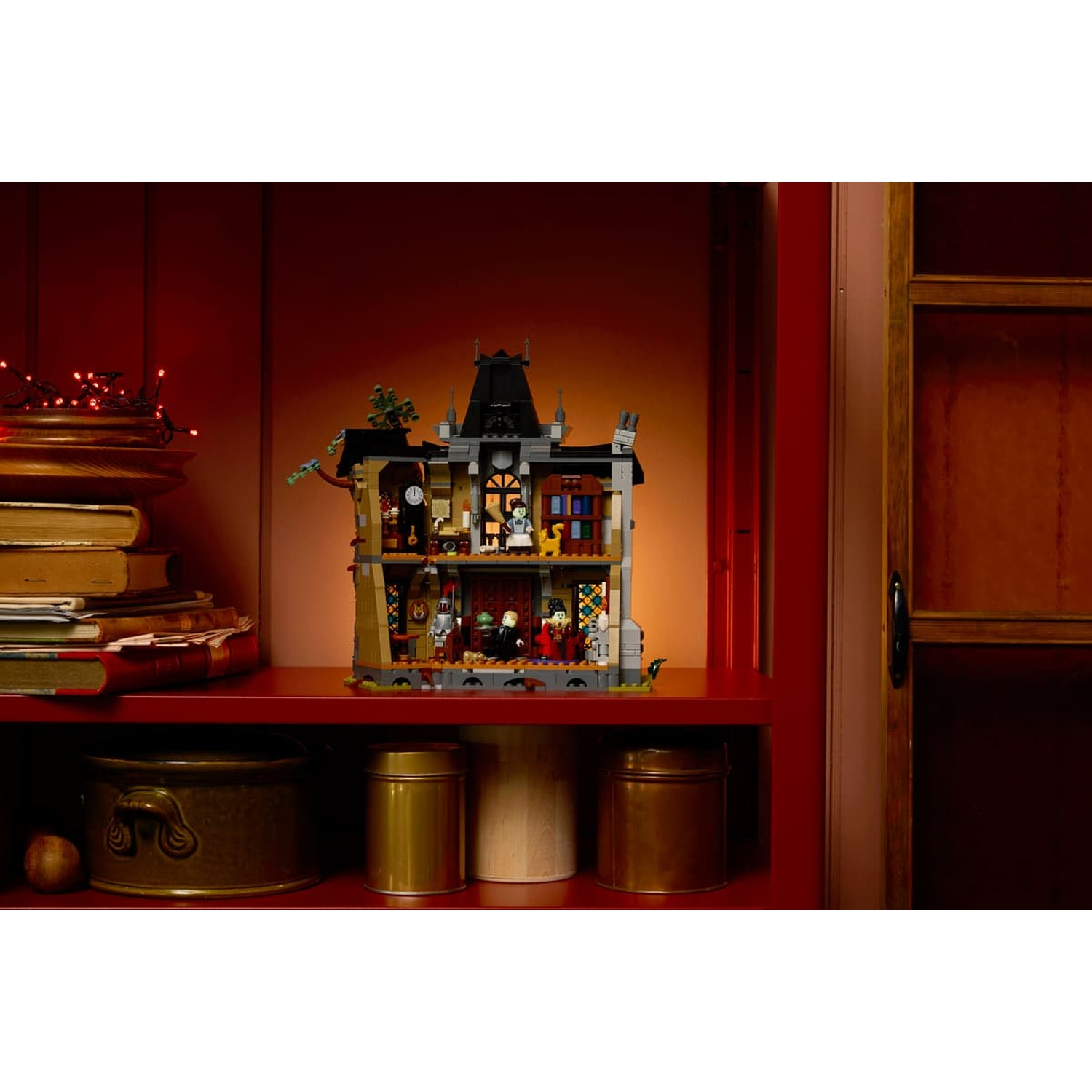 An illuminated Lego haunted house sits on a red shelf beside books and metal canisters.