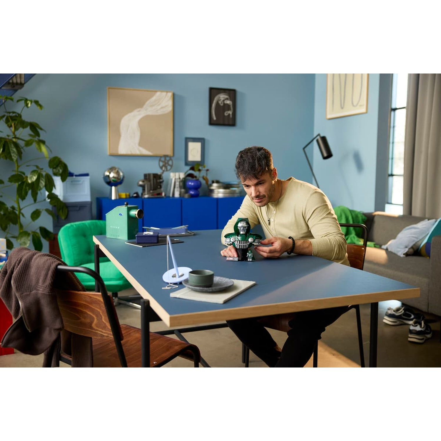 Un homme est assis à une table bleue dans un salon, assemblant un petit modèle vert.