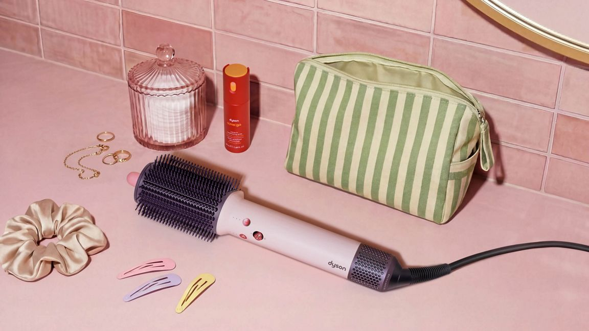 Eine Dyson-Haarbürste, eine Kosmetiktasche, Schmuck und Haarschmuck liegen auf einer rosa Ablage vor rosa Fliesen.