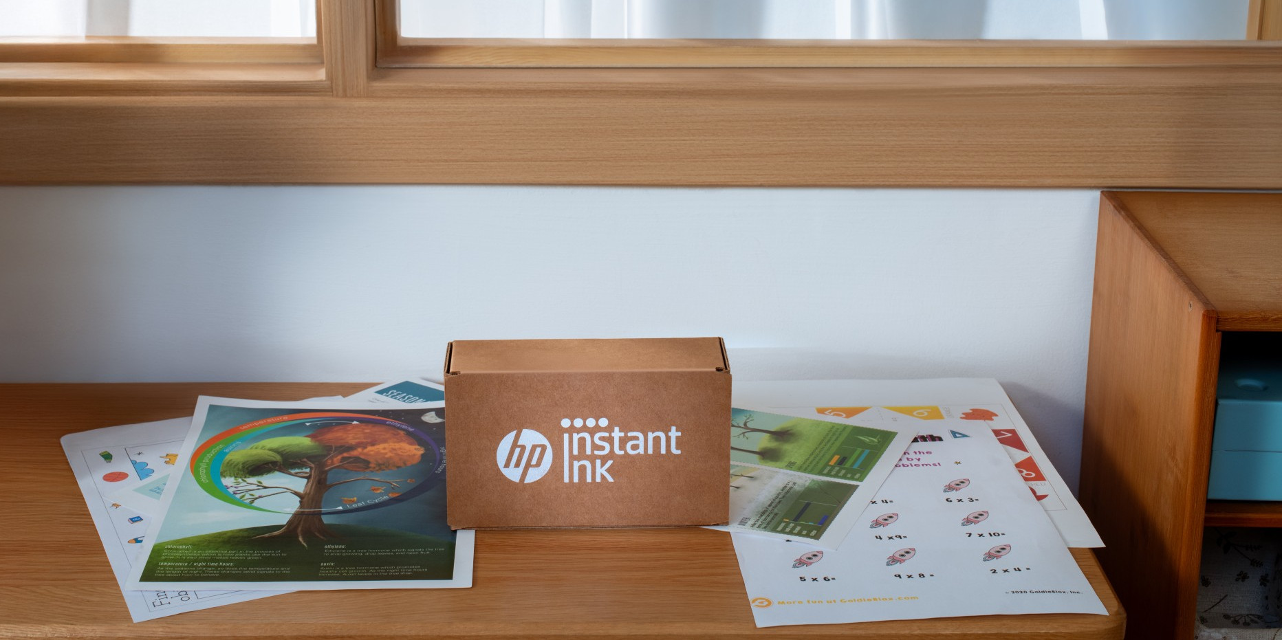 Karton HP Instant Ink na drewnianym biurku w otoczeniu wydrukowanych arkuszy edukacyjnych na tle białej ściany.
