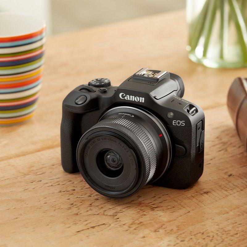 Eine schwarze Canon EOS Kamera auf einem Holztisch, daneben eine gestreifte Tasse.
