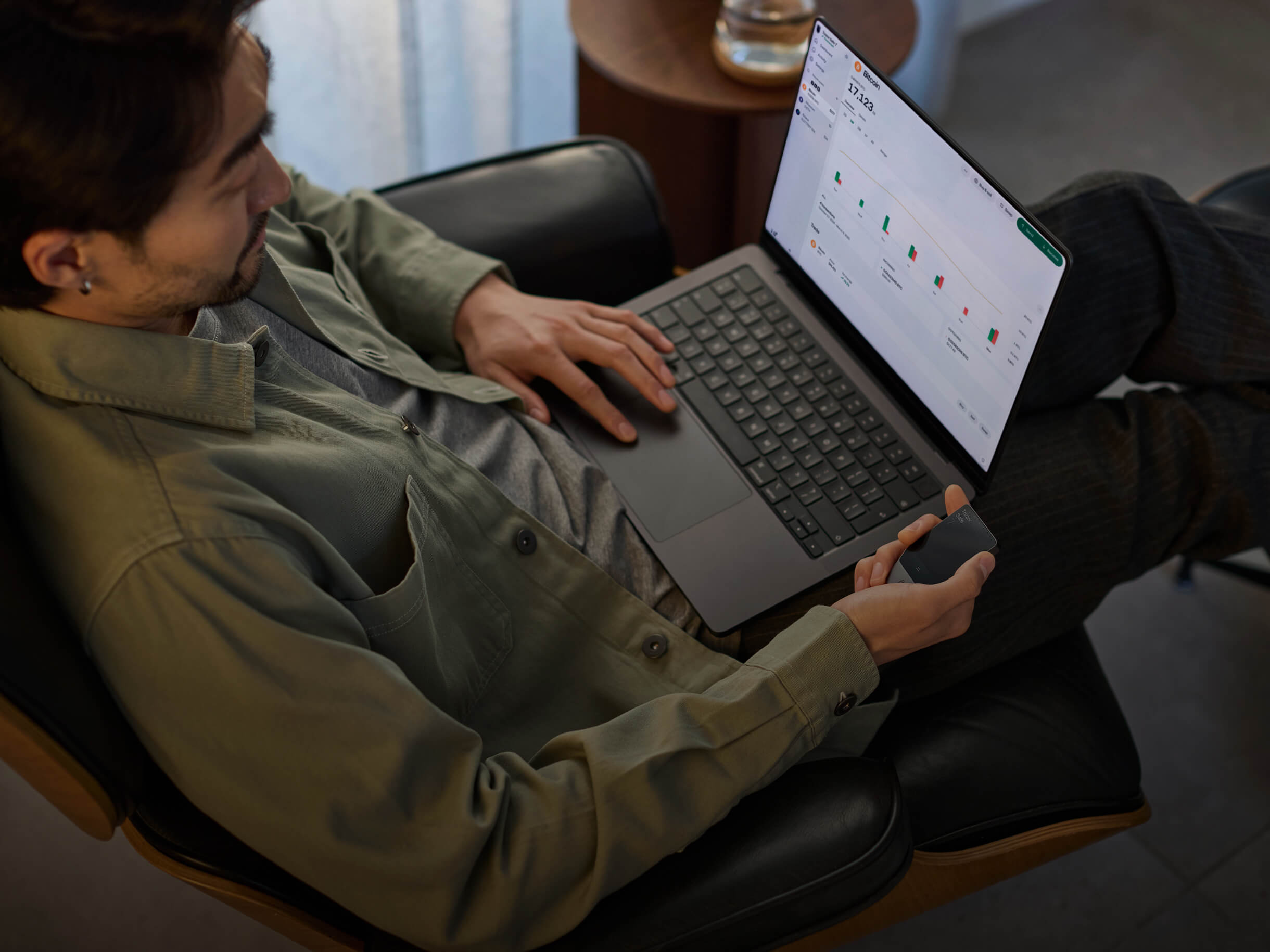 Ein Mann sitzt in einem schwarzen Sessel, benutzt einen Laptop mit Krypto-Charts und hält ein kleines Gadget in der Hand.