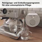 Eine silberne Espressomaschine mit Reinigungs- und Entkalkungsprogramm, vor einer gefliesten Wand.