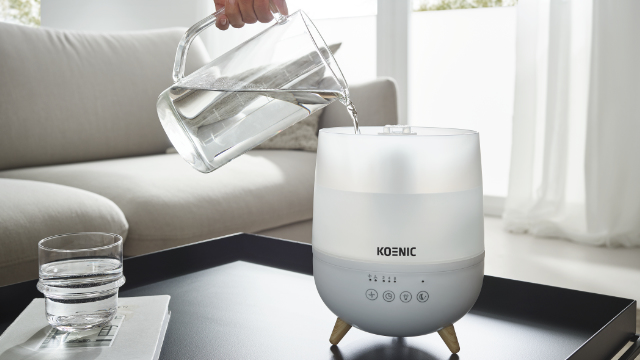 Une main verse de l'eau d'une cruche en verre dans un humidificateur blanc moderne sur une table noire.