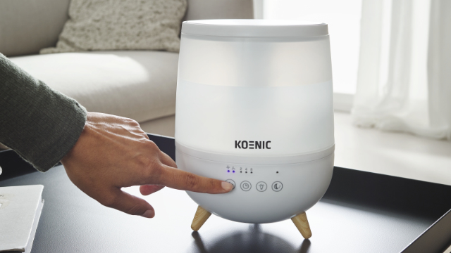 Une main actionne un humidificateur blanc Koenic sur une table sombre.