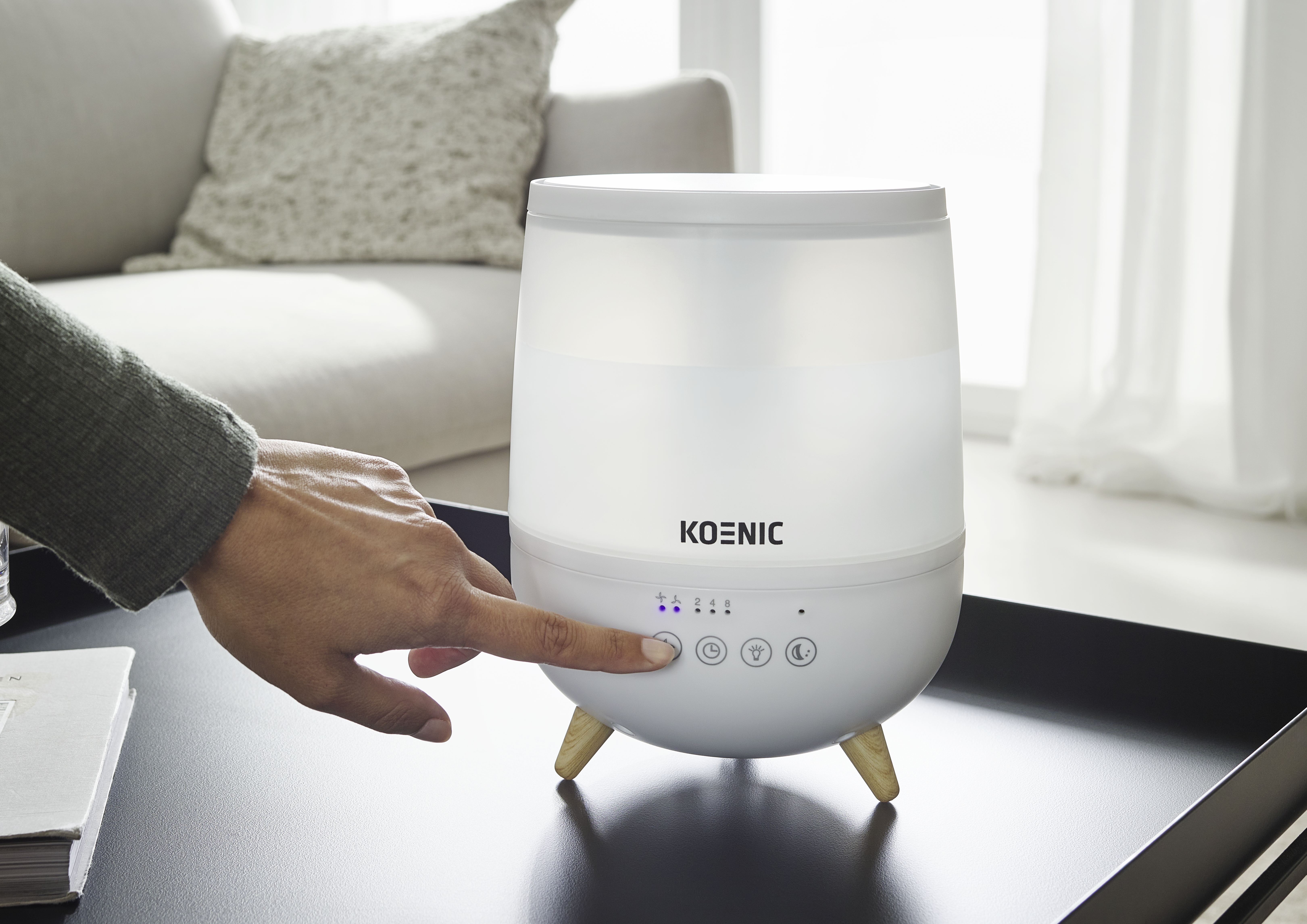 Une main appuie sur la commande tactile d'un humidificateur blanc Koenic sur une table sombre dans une pièce lumineuse.