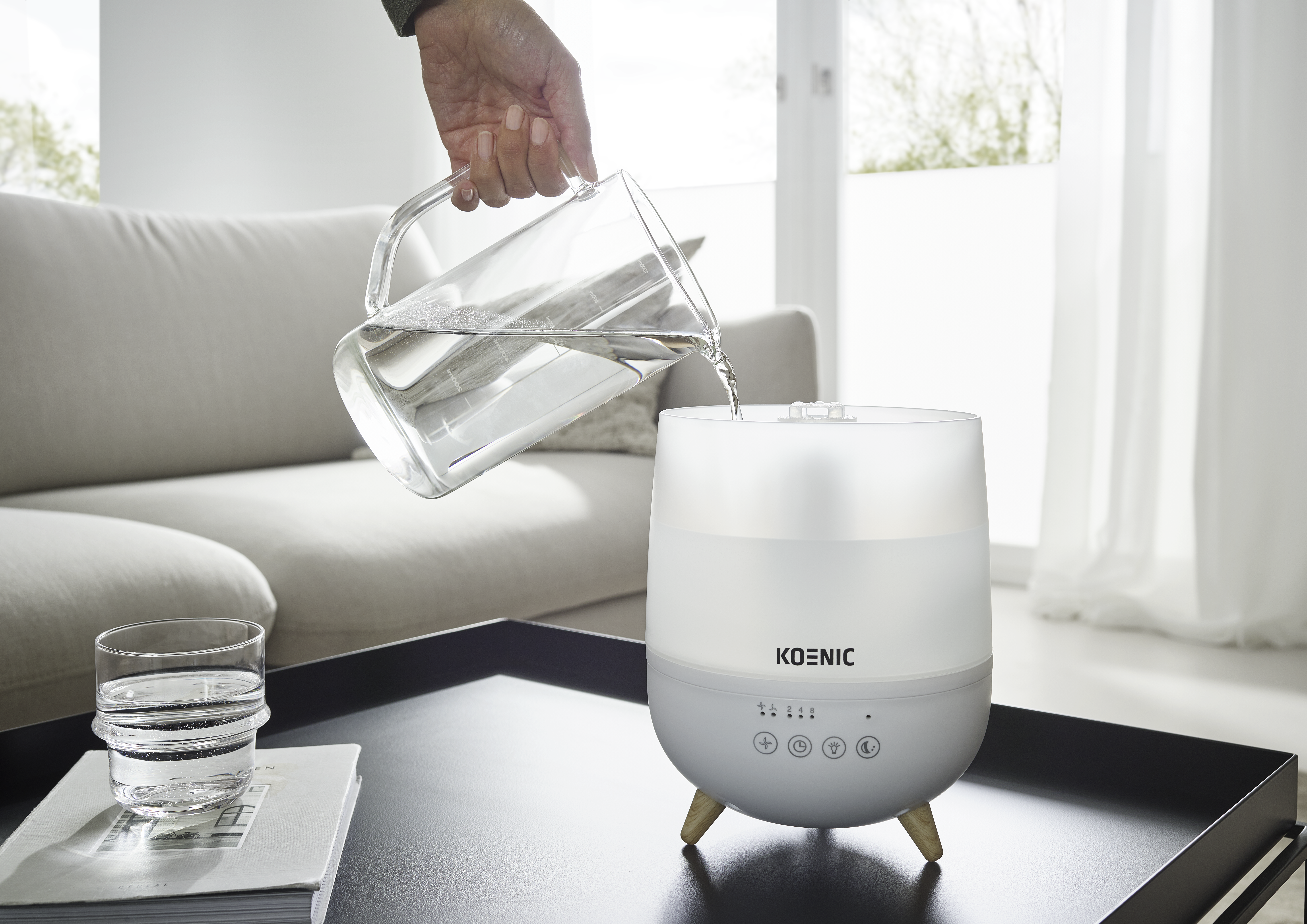 Une main verse de l'eau d'une carafe dans un diffuseur blanc sur une table avec un livre et un verre.