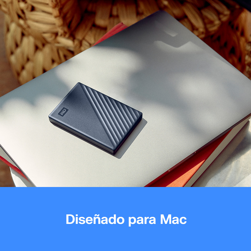 Un disco duro externo azul descansa sobre un portátil plateado, con un libro rojo y una cesta tejida debajo.